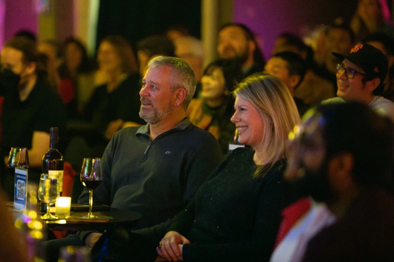 Audience enjoying a performance, with a middle-aged couple visible, seated at a table with wine and a candle, surrounded by other spectators in a dimly lit setting.