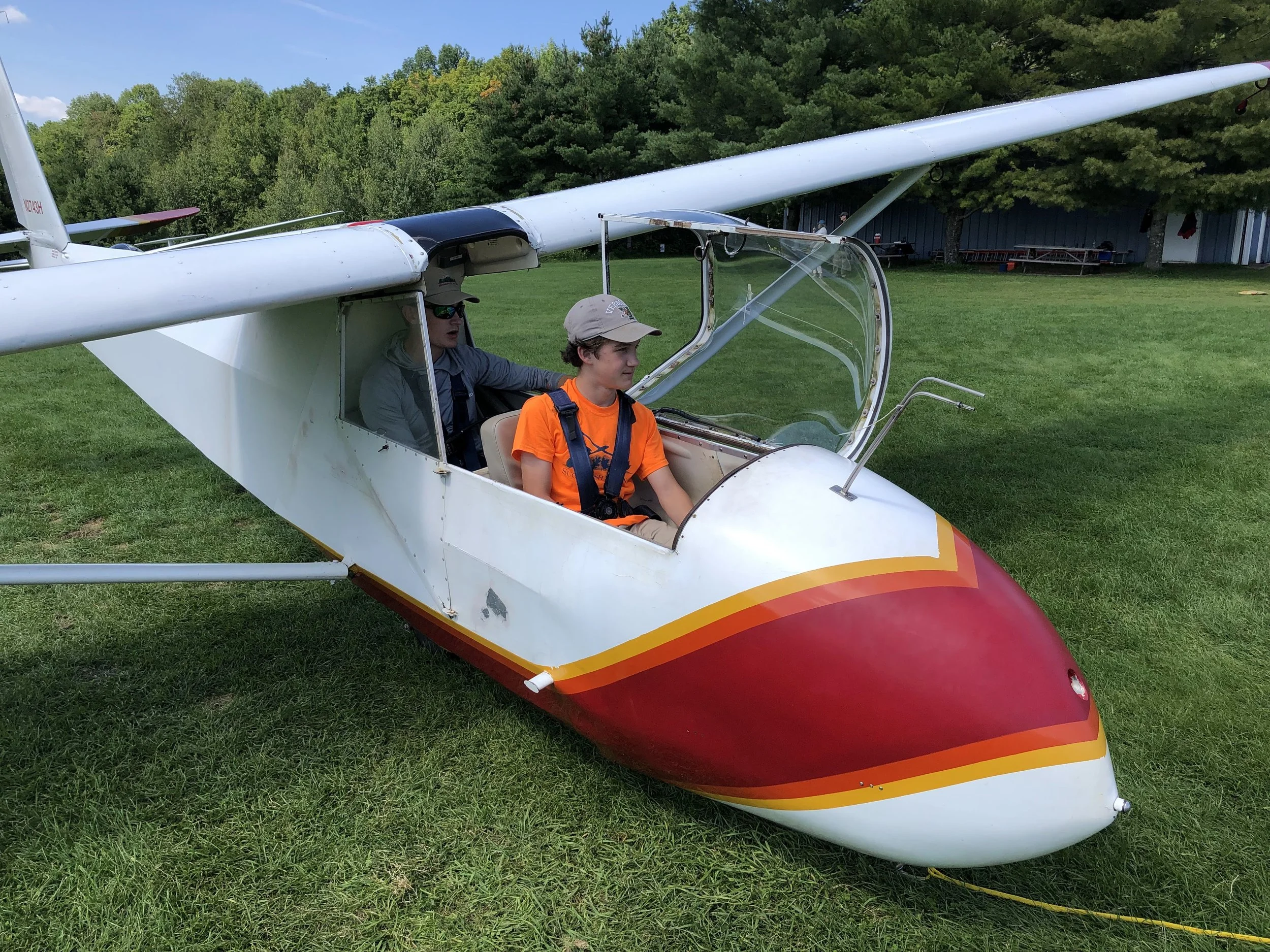 A small white glider plane with red and yellow accents on grassy ground, with two young boys inside, one in the pilot seat and the other in the co-pilot seat, surrounded by a green tree landscape.