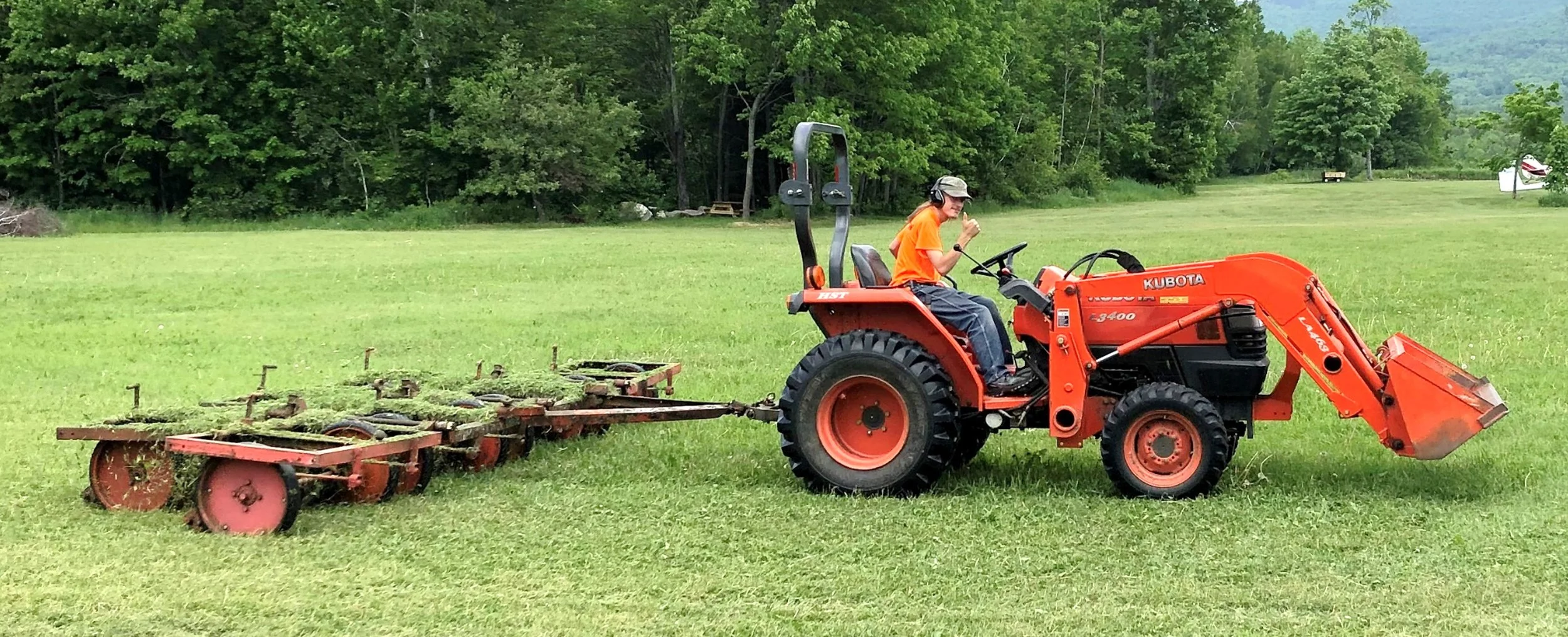 A person in an orange shirt and cap driving a red Kubota tractor on a grassy field, with a large wooded area in the background.