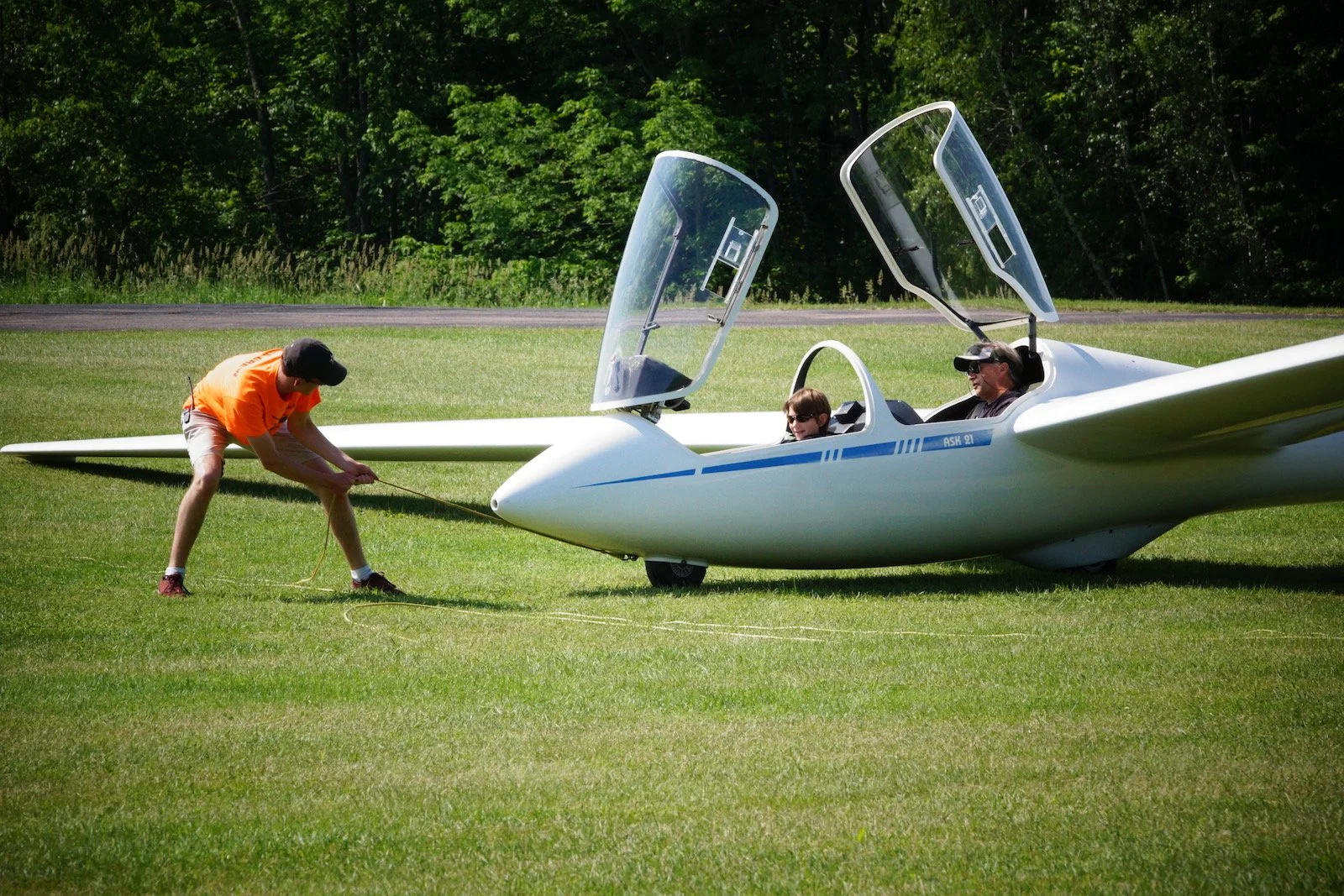 A man in an orange shirt and shorts is tying down a white glider aircraft, while two people, including a child, are sitting inside the cockpit with open canopies, on a grassy field with trees in the background.