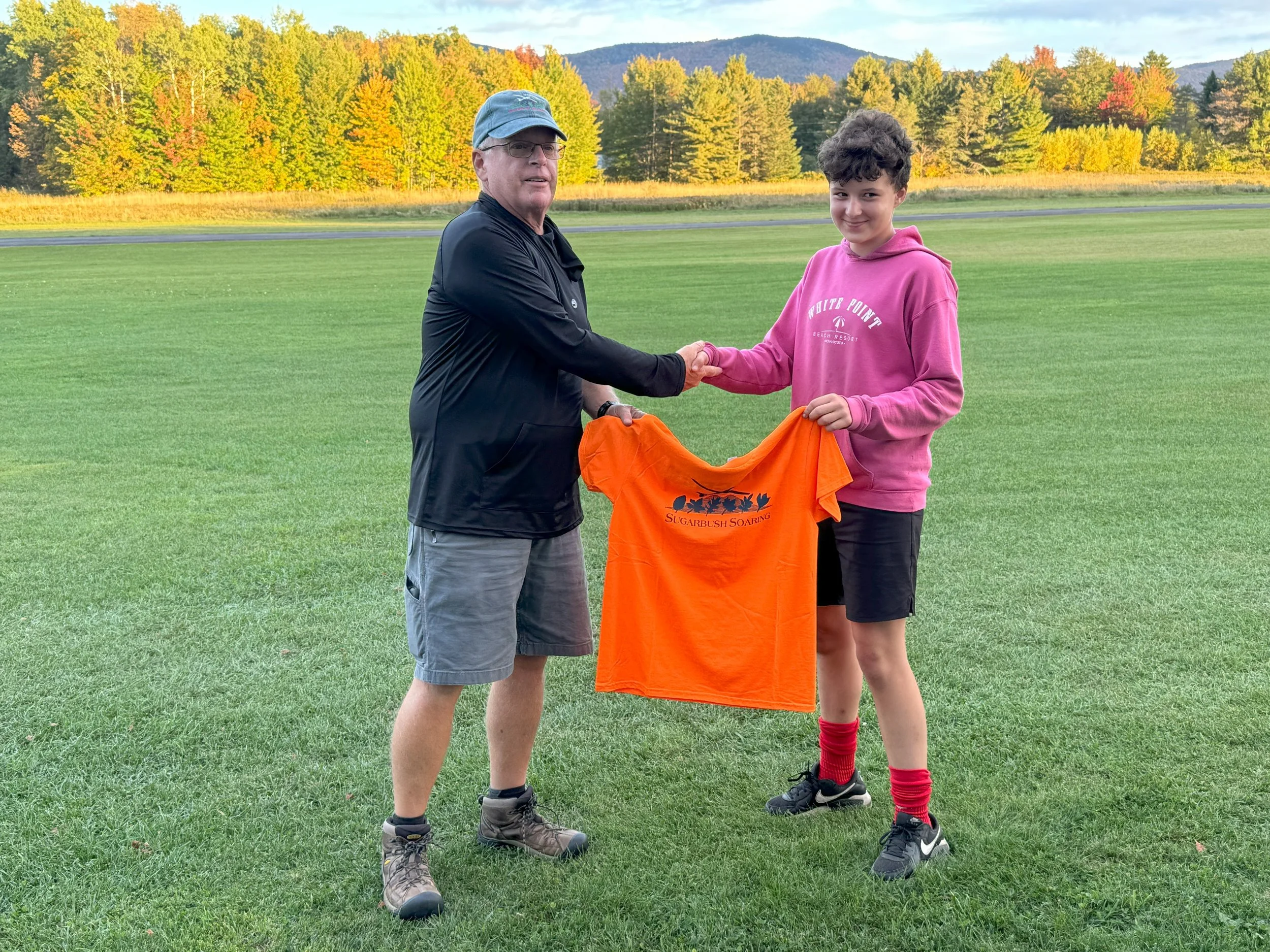 A man with dark, curly hair and a beard, wearing clear glasses and a black sweater, sitting indoors with a neutral expression.