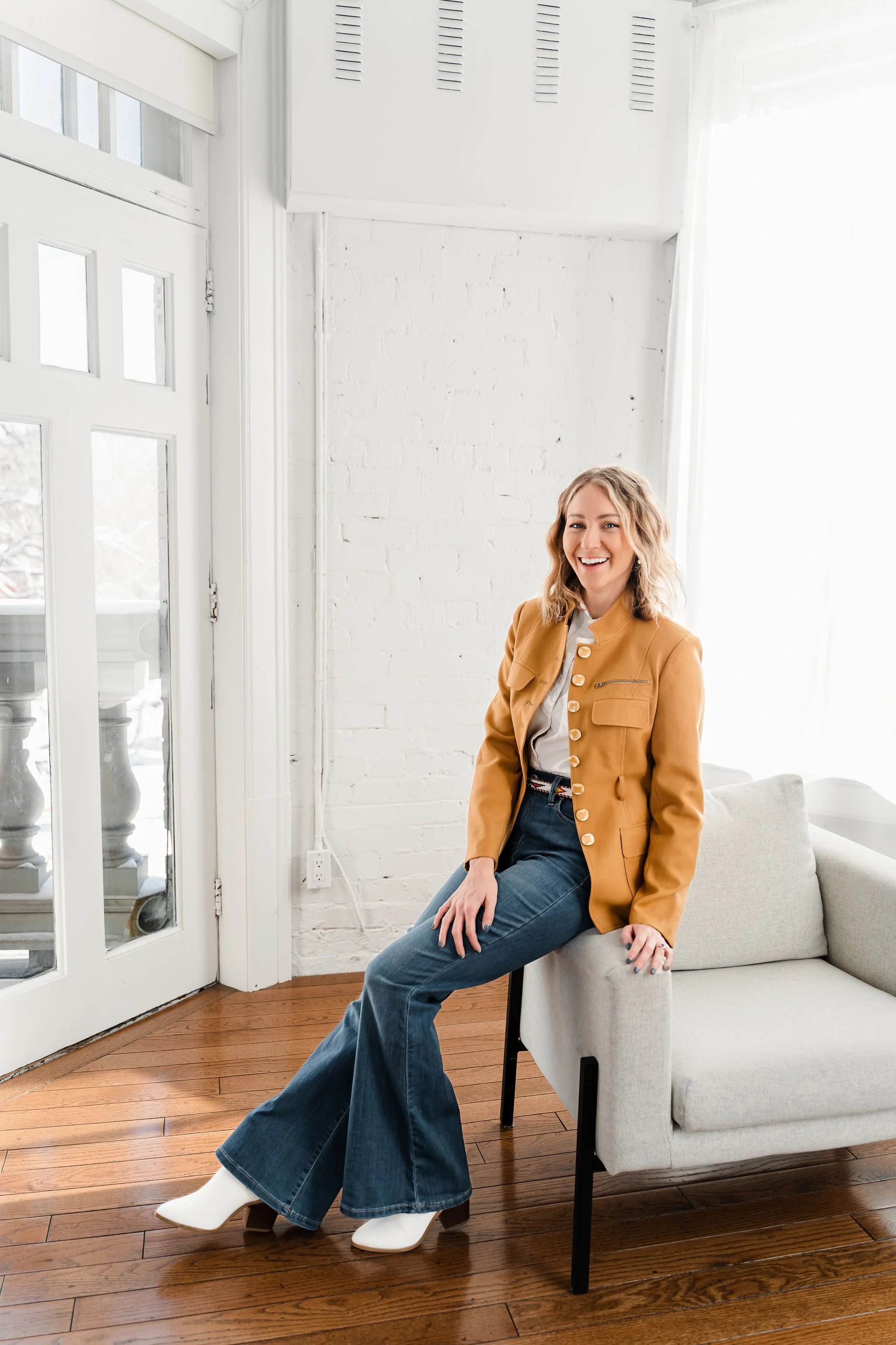Person sitting on a modern light-colored chair in a bright room, wearing a mustard blazer, blue jeans, and white boots.