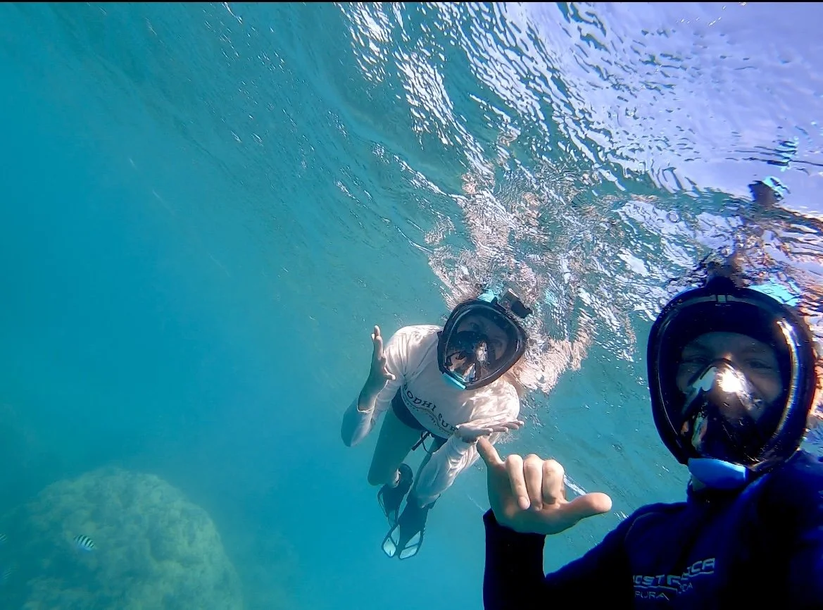 Two people snorkeling underwater with full-face masks, clear blue water, one person making a hand gesture.