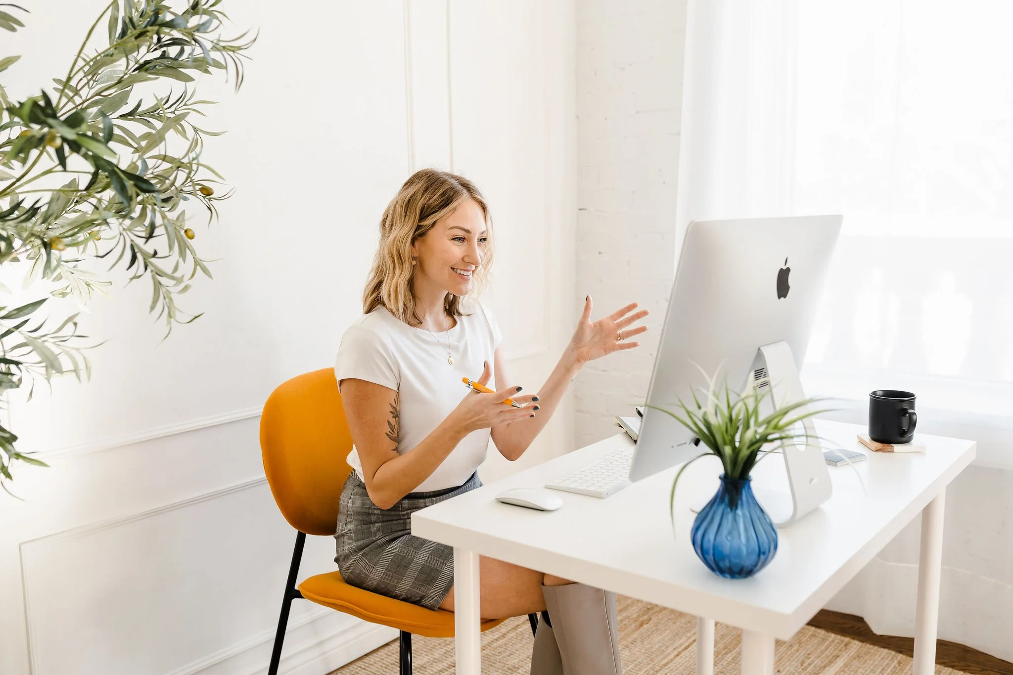 Woman gesturing during a video call on a computer in a bright office with plants and a mug on the desk.
