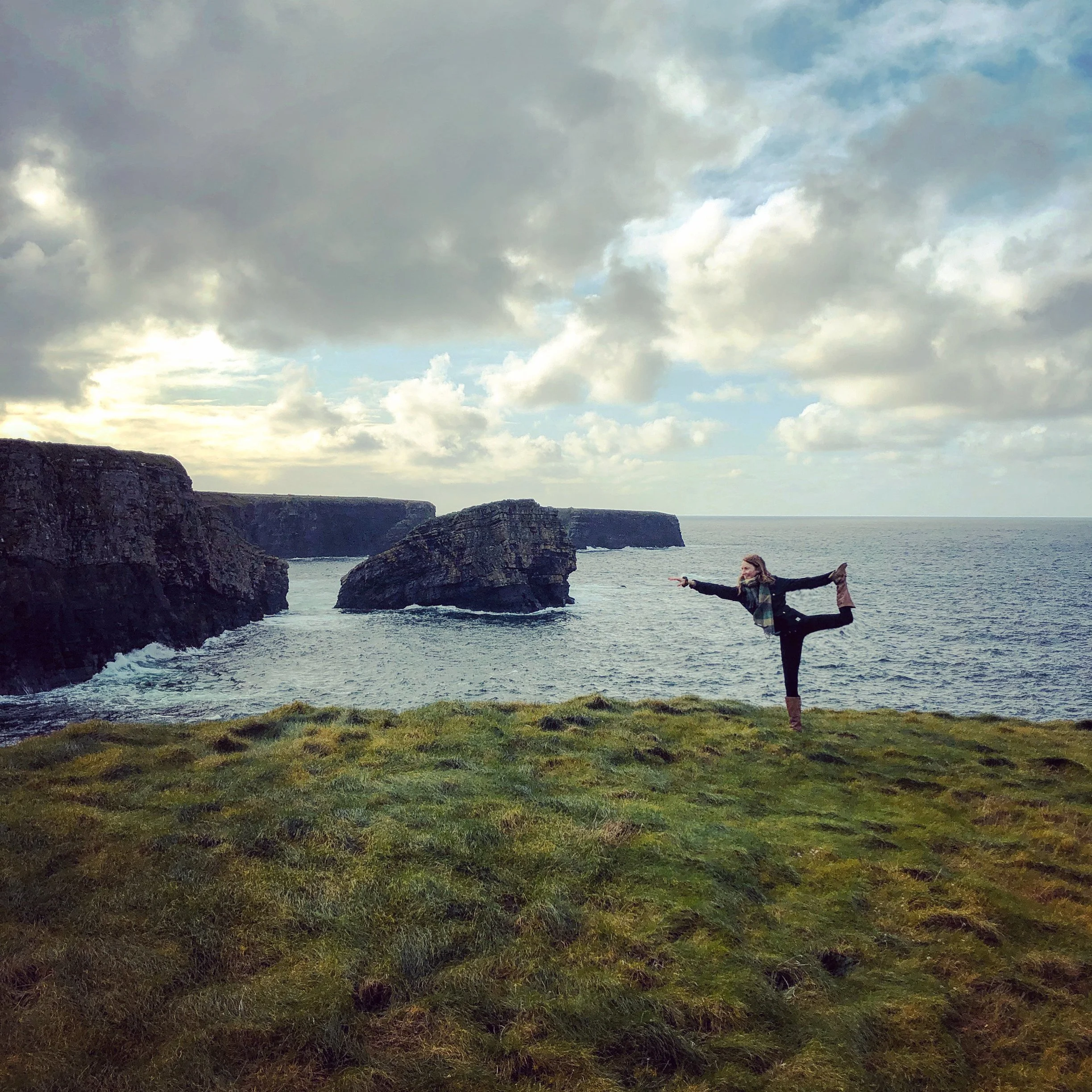 Person doing yoga pose on grassy cliff overlooking rocks and ocean under cloudy sky