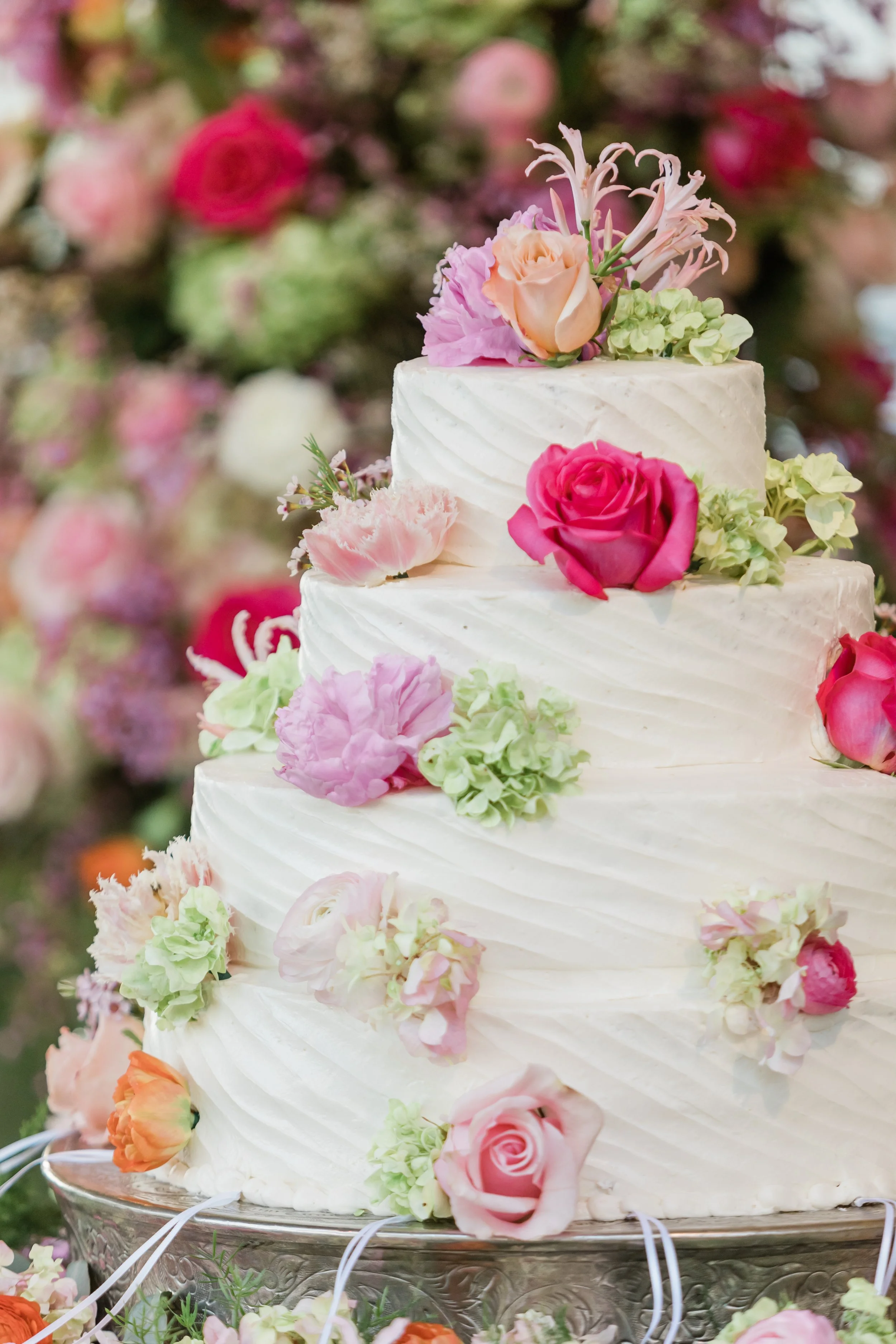 A multi-tiered white wedding cake decorated with various pink, peach, and green flowers, including roses and hydrangeas, on a silver cake stand.
