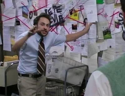 Charlie Kelly from It’s Always Sunny in Philadelphia with a crazed look on his face standing in front of a corkboard covered with papers connected with chaotic red strings