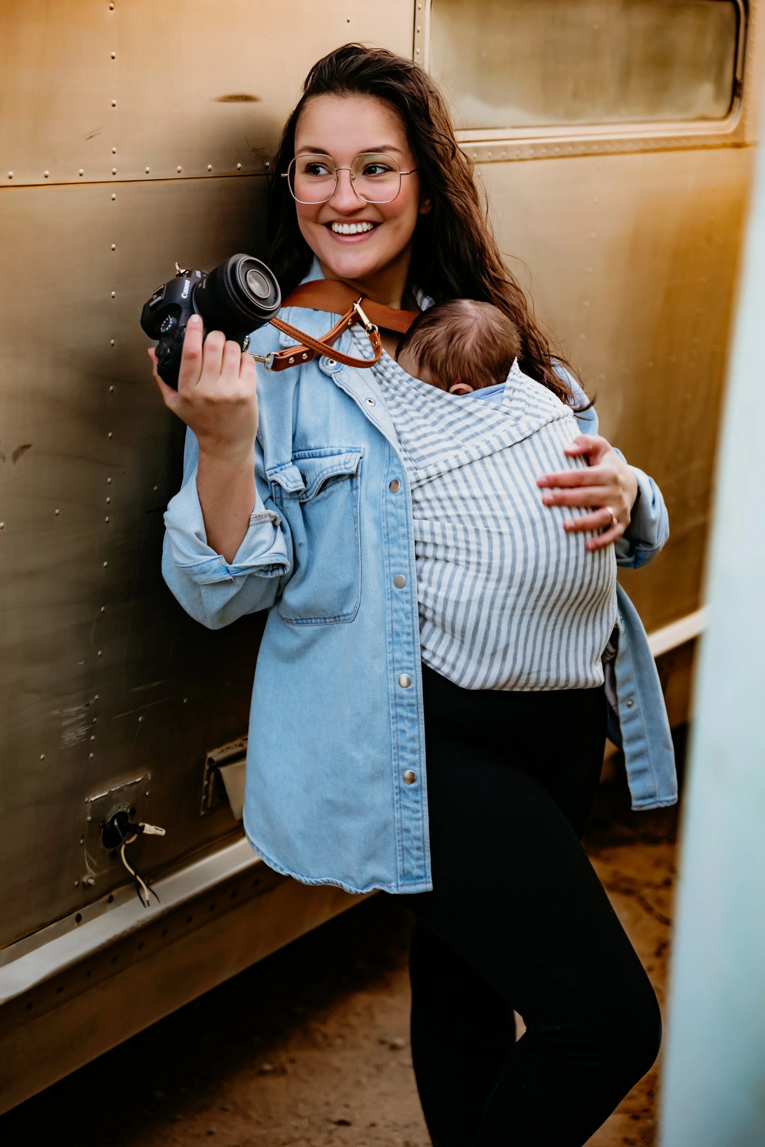 A woman with long brown hair wearing glasses, a denim jacket, and a striped baby carrier holding a sleeping baby in front of a wooden wall.