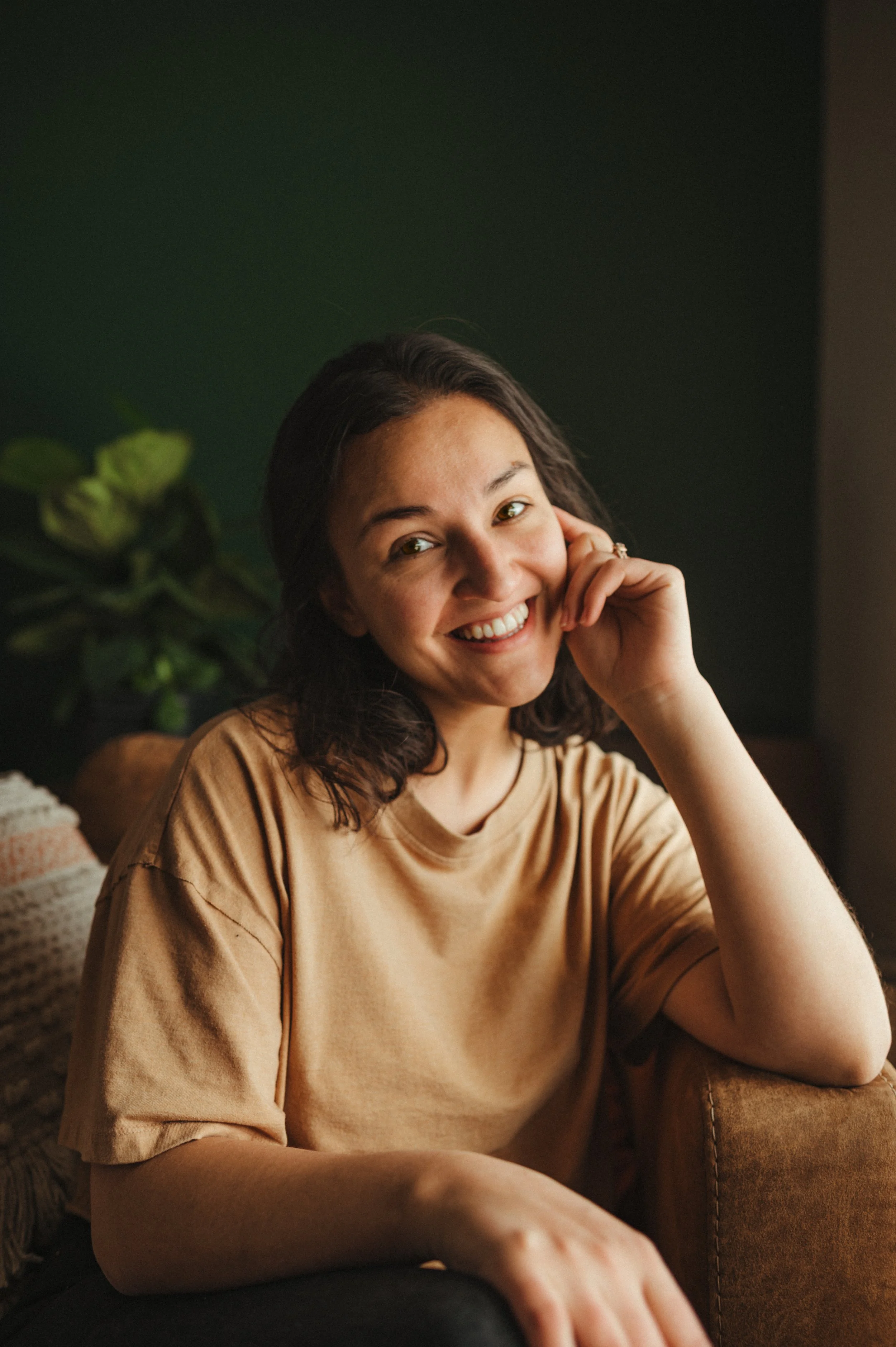 A smiling woman with dark hair sitting on a brown sofa, wearing a tan t-shirt, with a plant and dark green wall in the background.