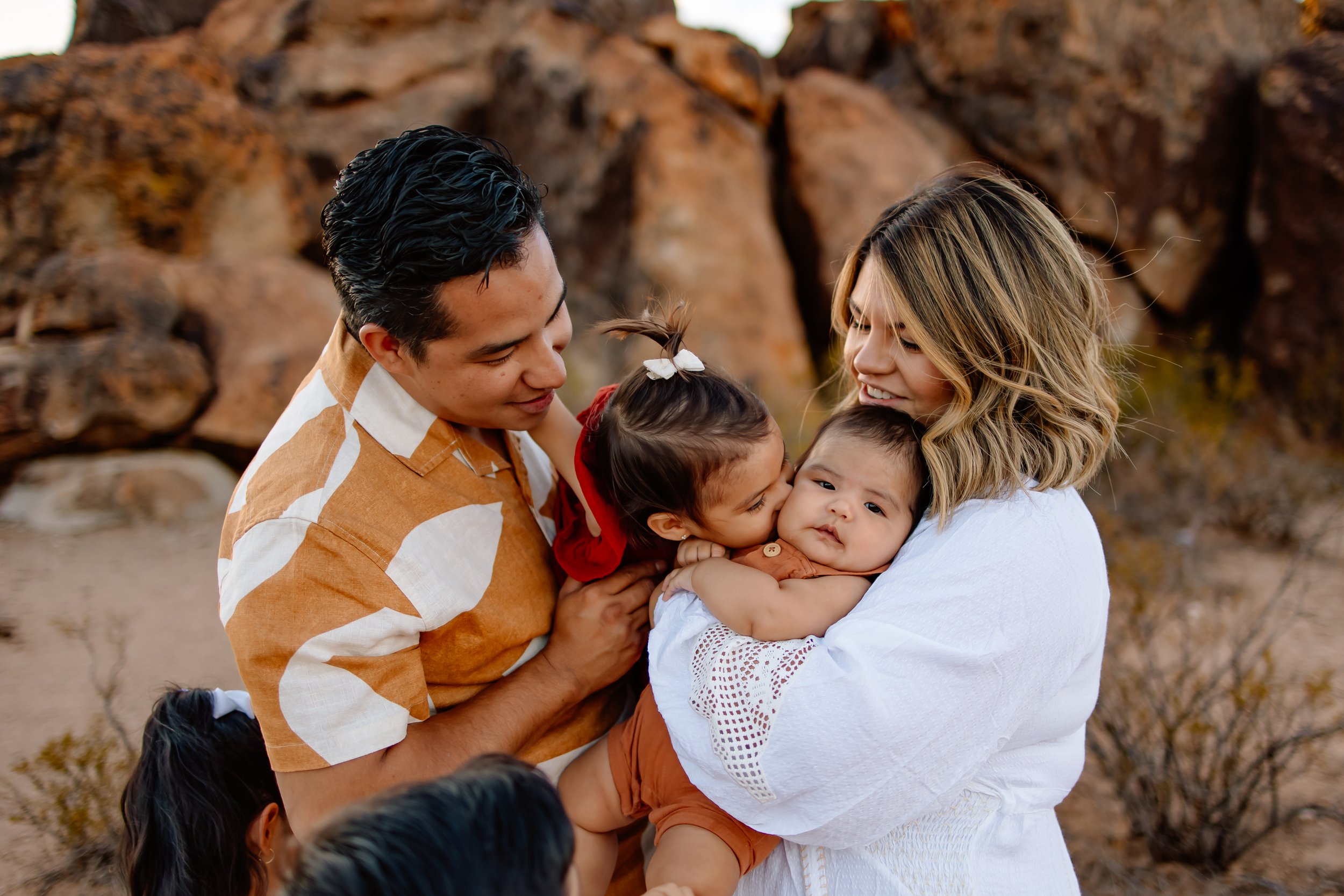 A family of five outdoors, with rocky terrain in the background. A woman is holding a baby, while a young girl kisses the baby on the cheek, and a man looks at them with affection.