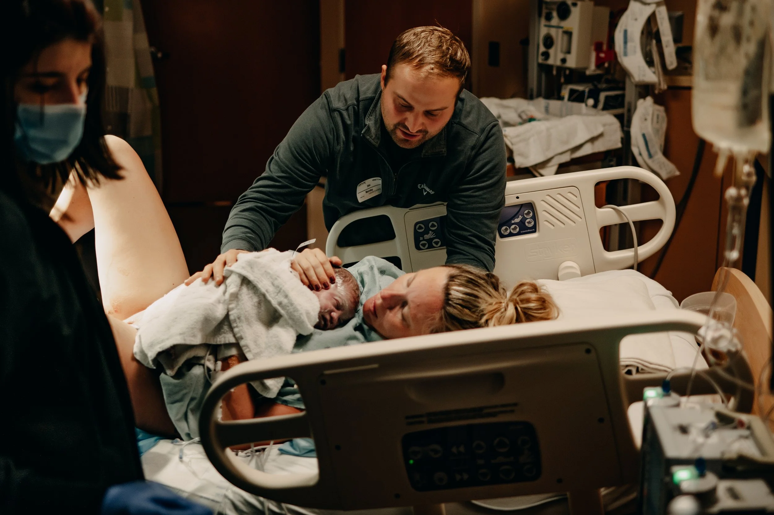 A woman in labor, lying in a hospital bed, is being attended by a nurse and a man, a doctor, who are helping her with the birth of a newborn.