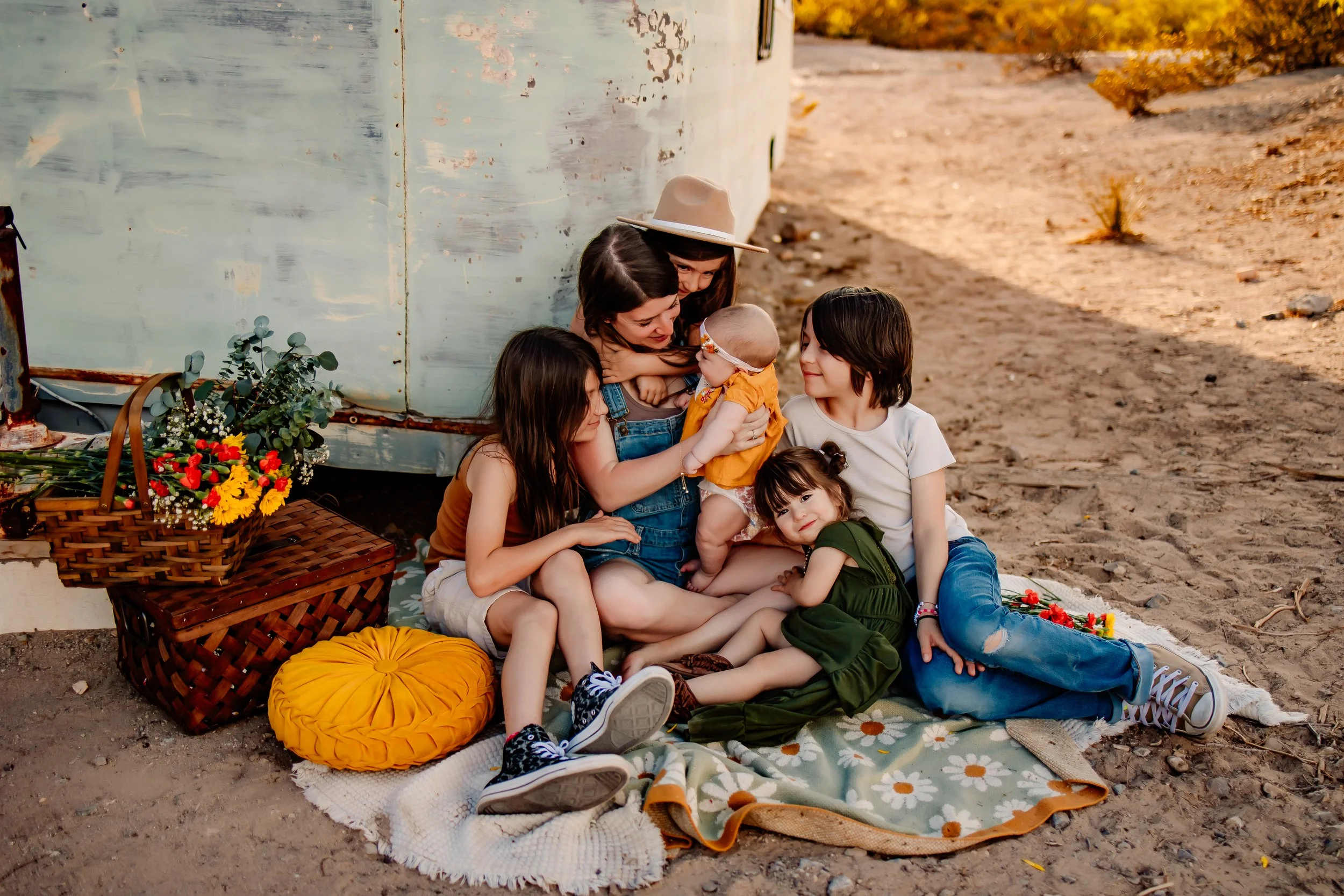 Family of five, mother, four children, sitting on a blanket outdoors near a distressed metal wall, with a basket of flowers and a yellow pillow, during sunset in a desert setting.