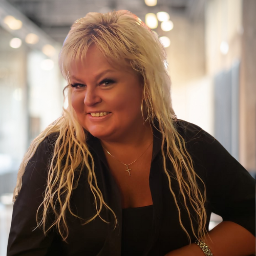 Smiling person with blonde hair in a black outfit indoors.