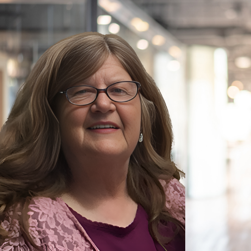 Smiling person with long, wavy hair and glasses, wearing a pink top and a maroon blouse, standing indoors with a blurred background of a corridor.