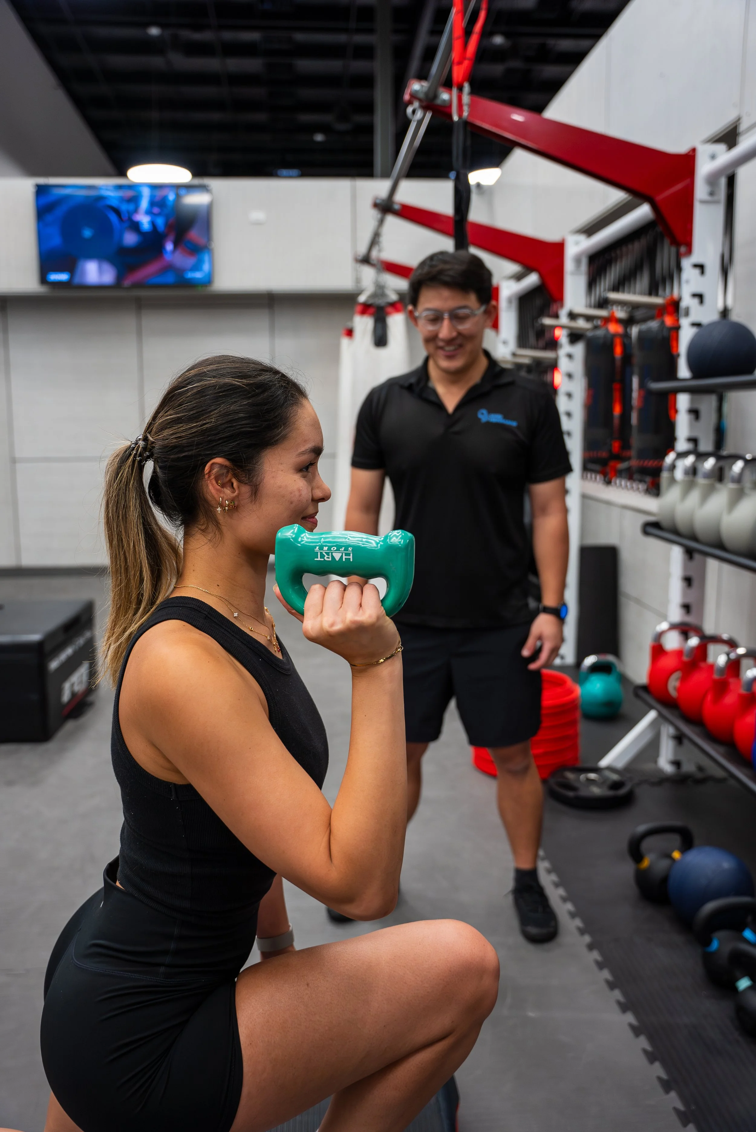 A woman is doing a goblet squat holding a green dumbbell in front of her chest, while a male trainer watches and encourages her in a gym with kettlebells and fitness equipment.