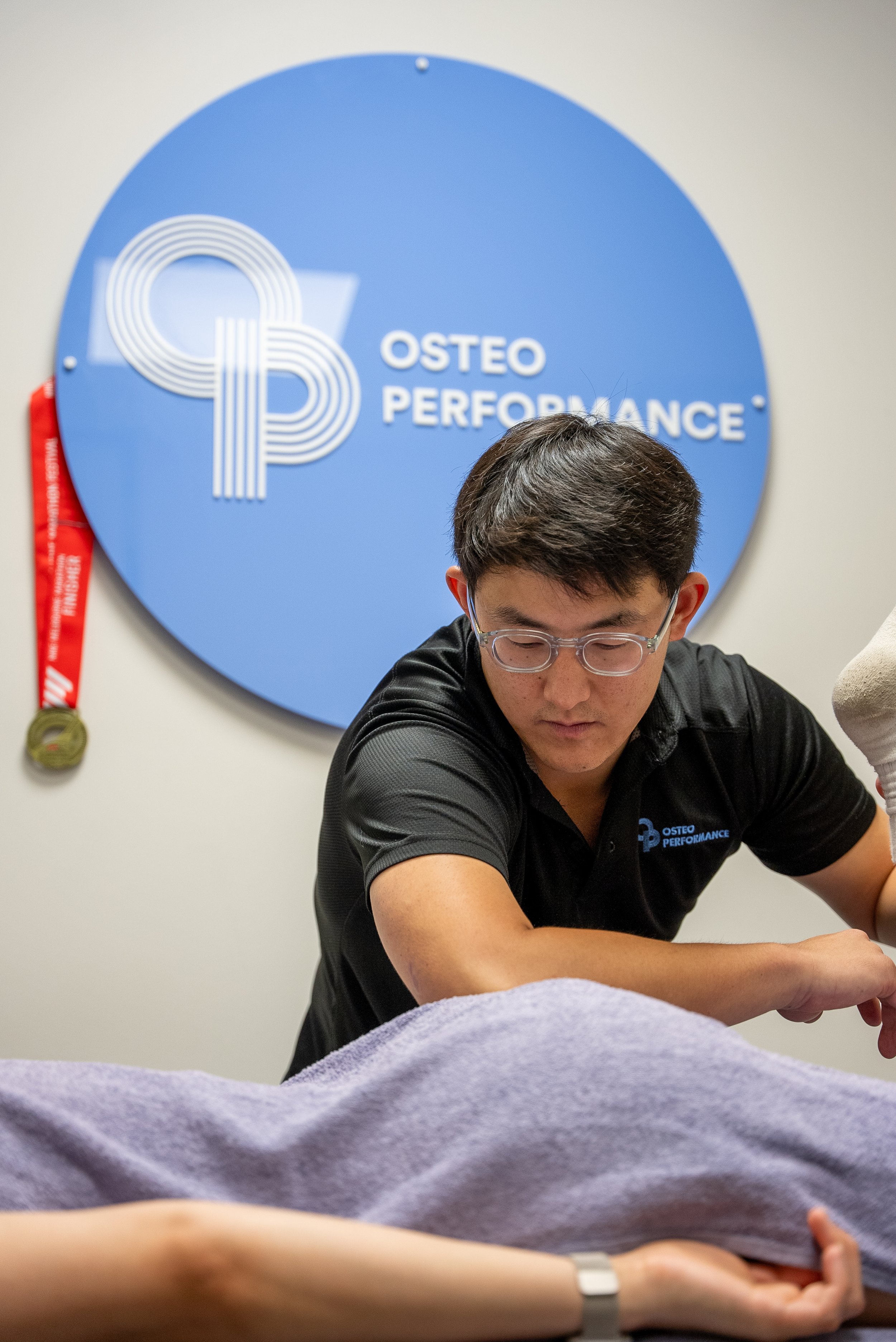 A man wearing glasses and a black polo shirt with 'Osteo Performance' logo performs a treatment on a client lying on a massage table. A blue sign with the logo and text 'Osteo Performance' hangs on the wall behind them.