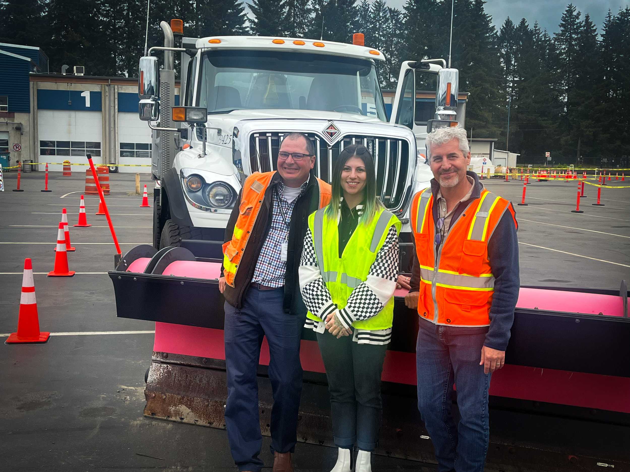 Three people wearing reflective safety vests standing in a parking lot with orange traffic cones, and a large snow plow truck in the background.
