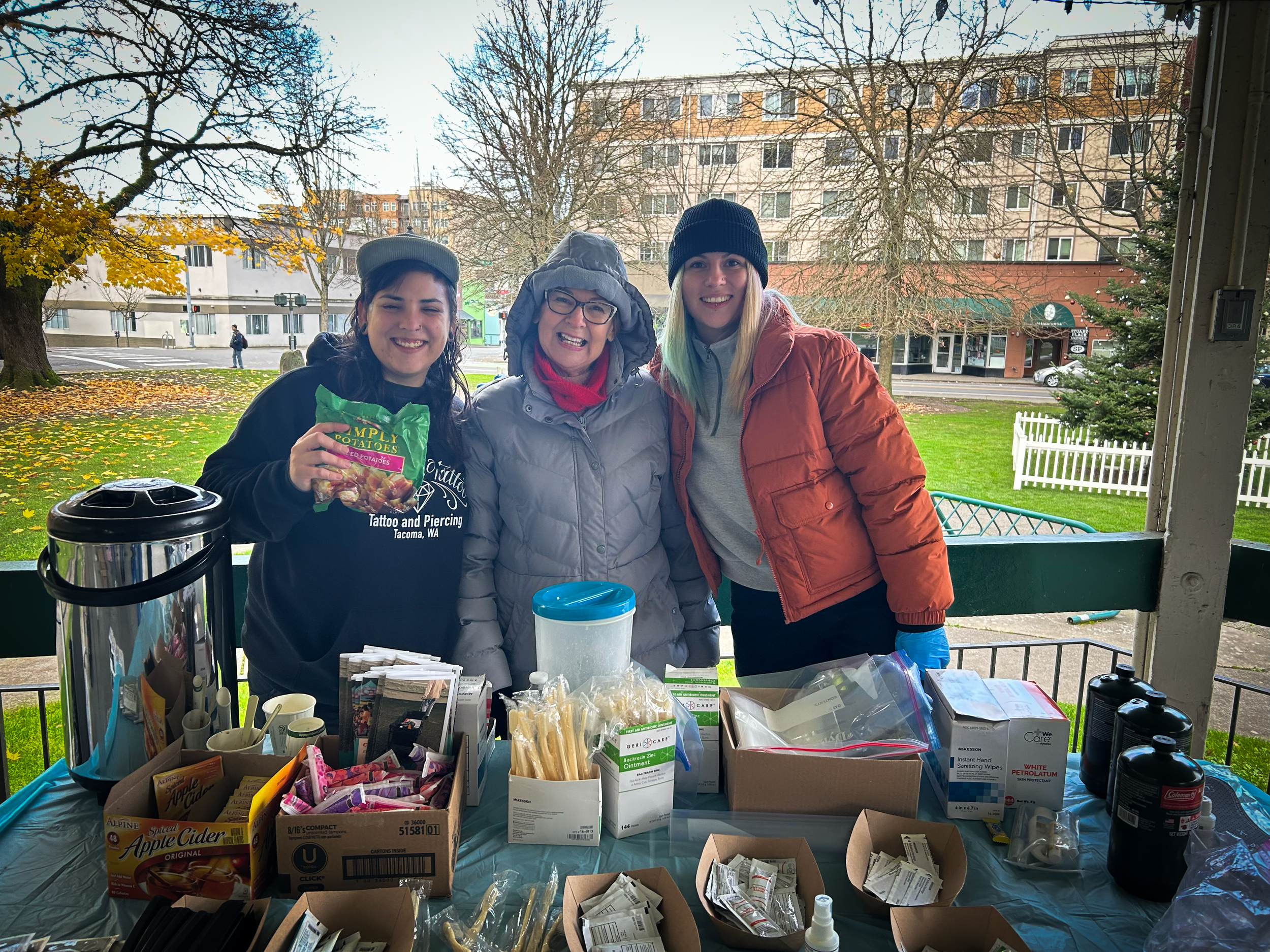 Three people standing behind a table with health supplies outdoors on a cloudy day, with trees and a building in the background.