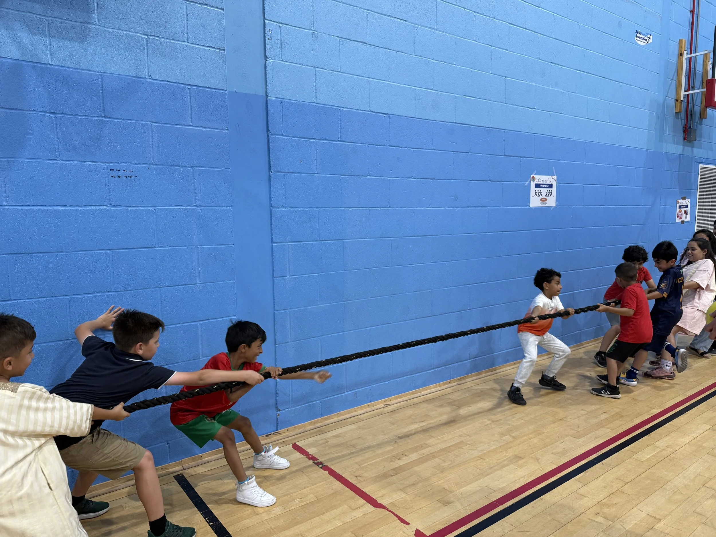 Children participating in a tug of war game inside a gymnasium with a blue wall.