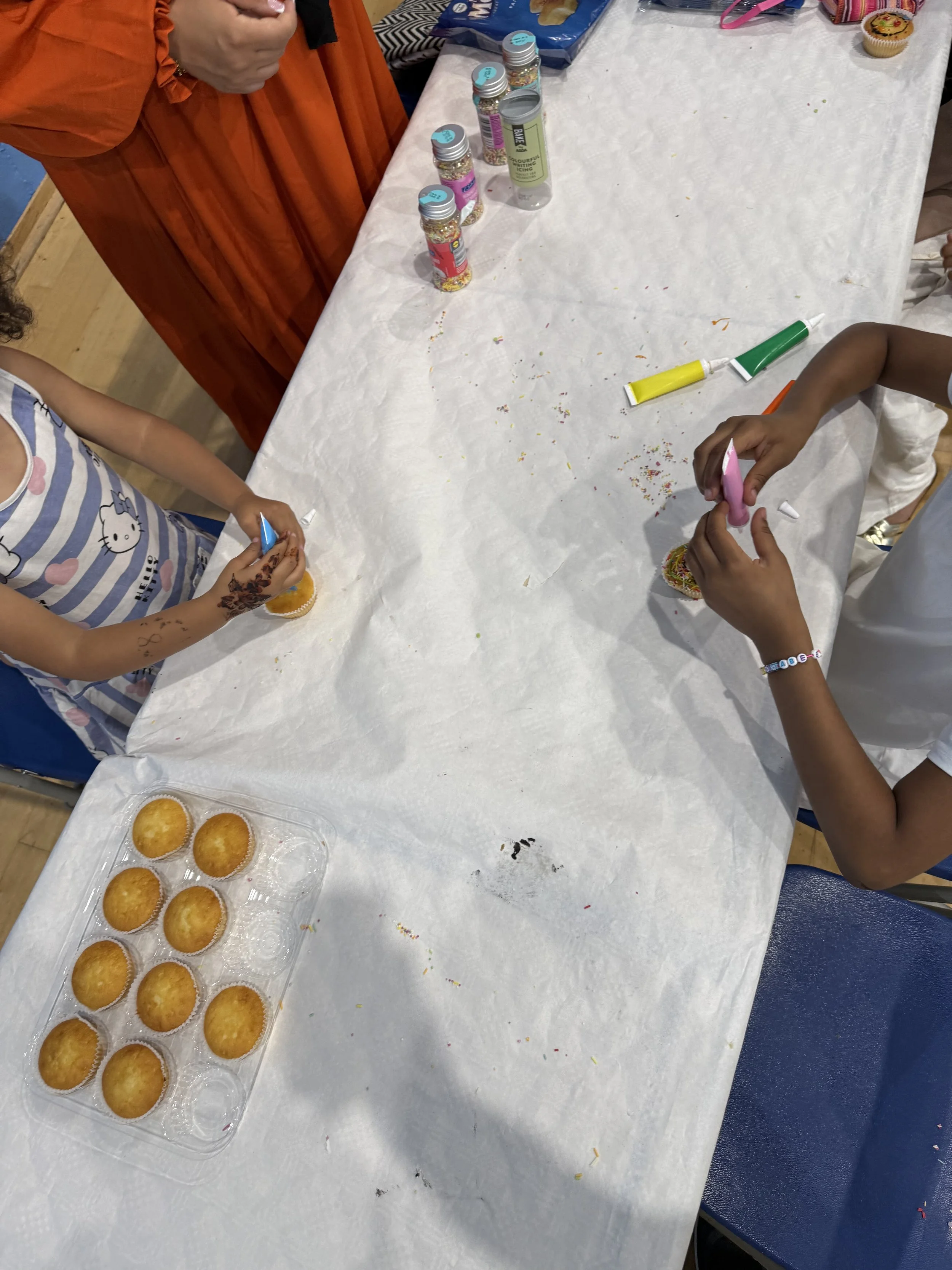 Children decorating cupcakes at a table with icing tubes and sprinkles, with a tray of plain cupcakes on the table.