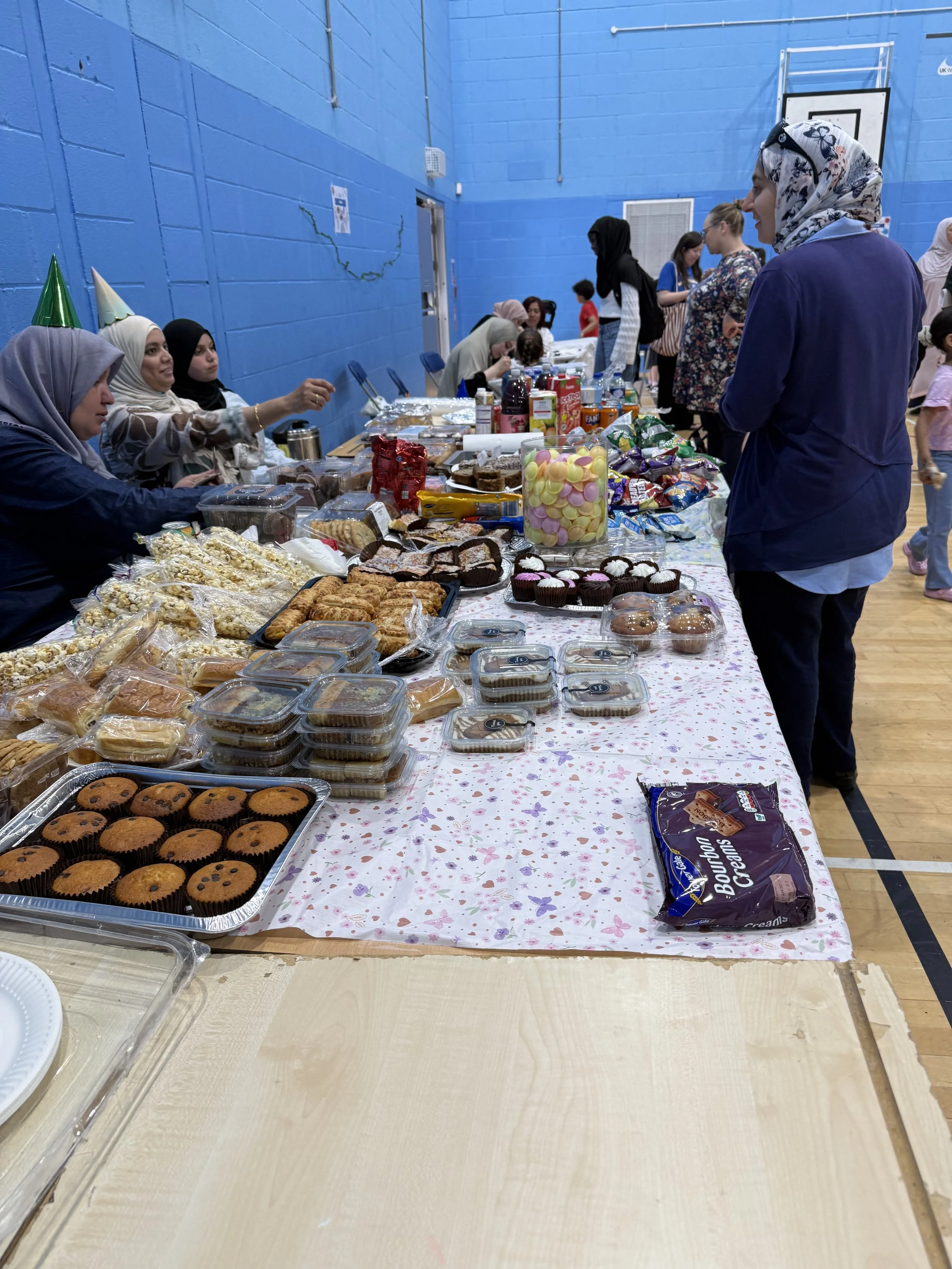 Table filled with various baked goods and snacks, in a gymnasium setting with people and blue walls