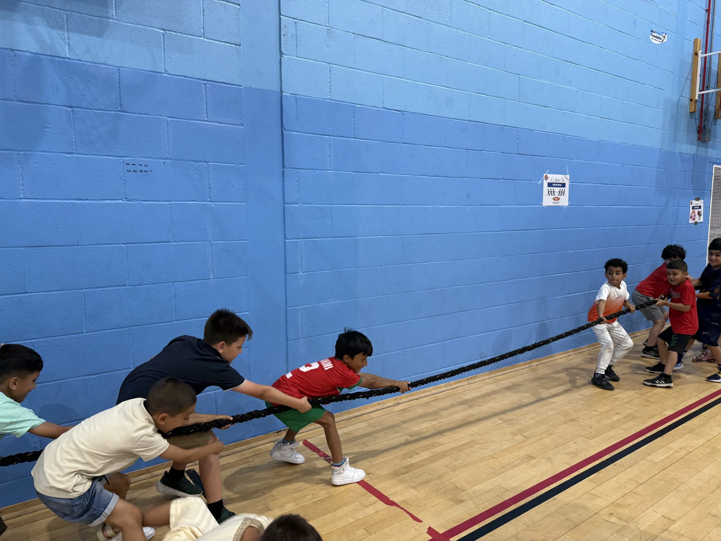 Children participating in a tug-of-war game inside a gymnasium with a blue wall and wooden floor, supervised by an adult.