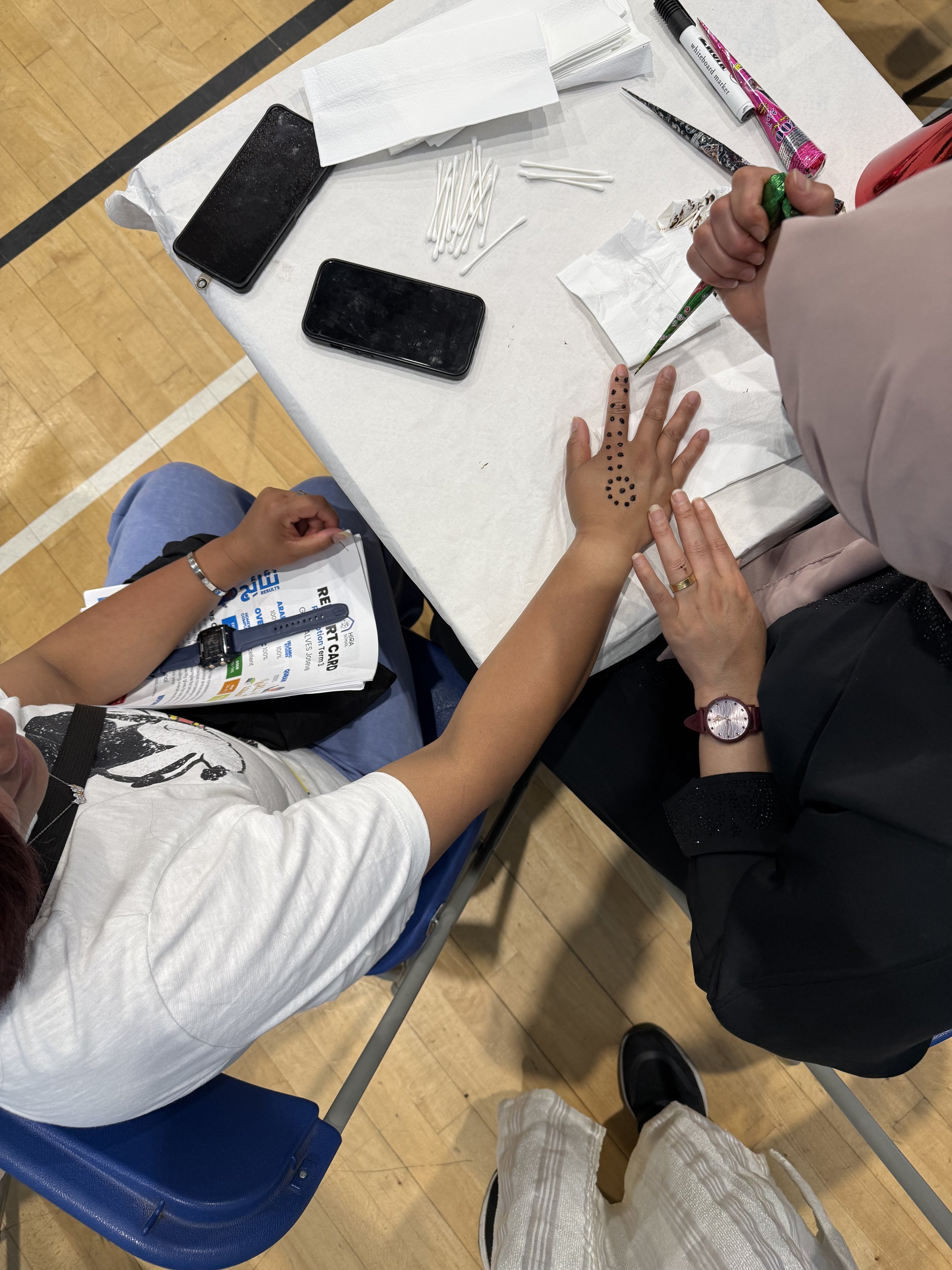 Health screening or blood donation setup at a community event or clinic, showing a person with their hand extended for testing or donation, surrounded by medical and personal items on a table, with another person assisting.
