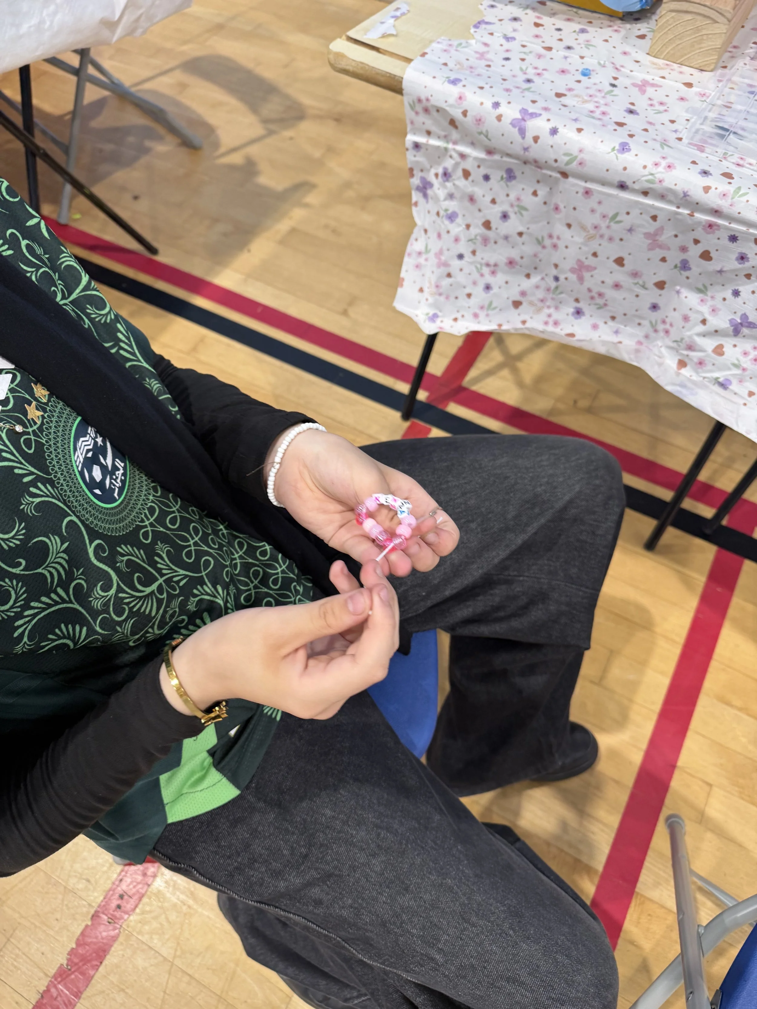 A person sitting in a gymnasium, holding a pink beaded friendship bracelet, with a table covered in a floral tablecloth in the background.
