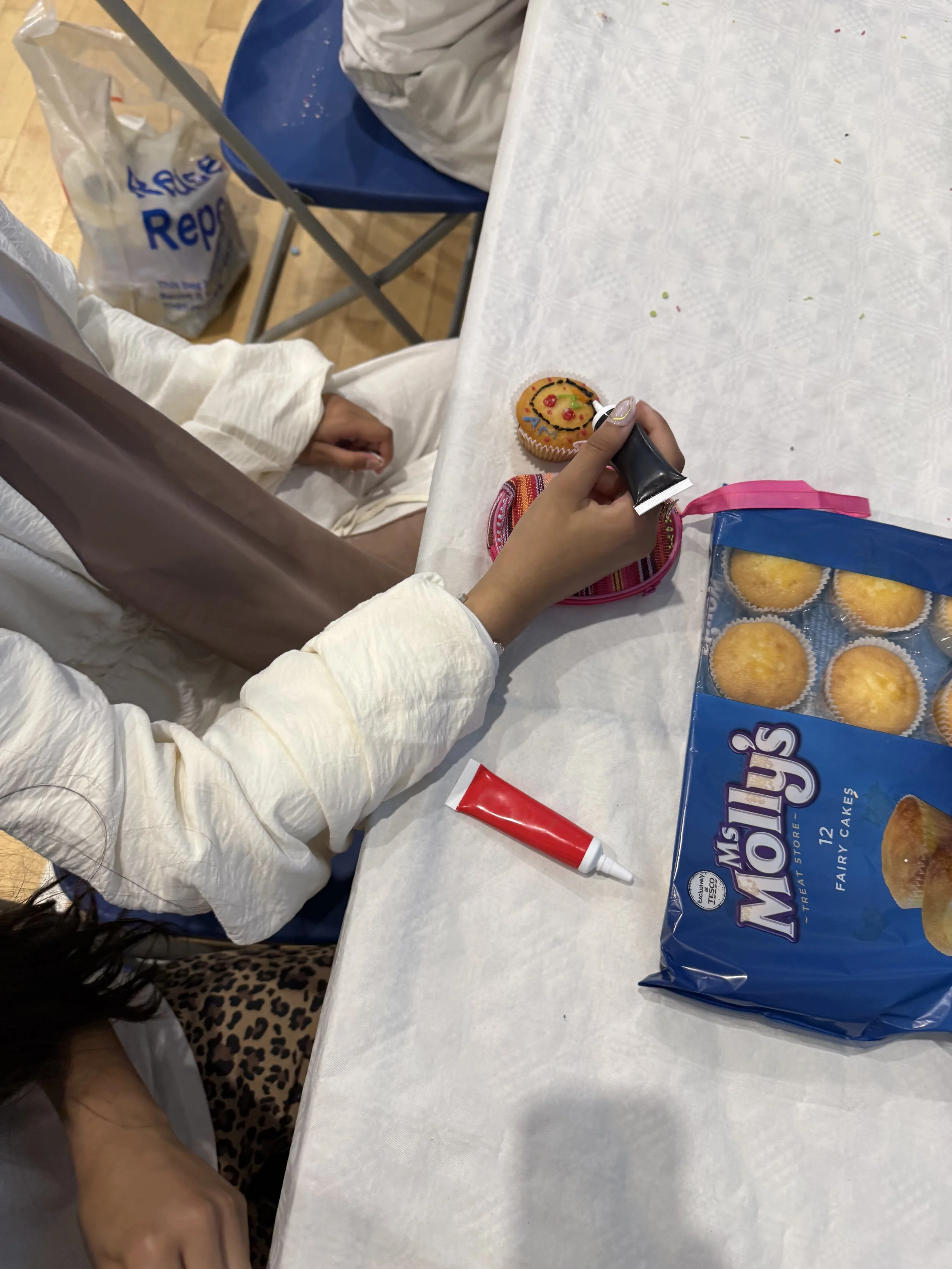 A white table with colorful sprinkles and a decorated cupcake being held with a piping tip. A package of Hostess cupcakes and some red and white frosting tubes are also on the table.
