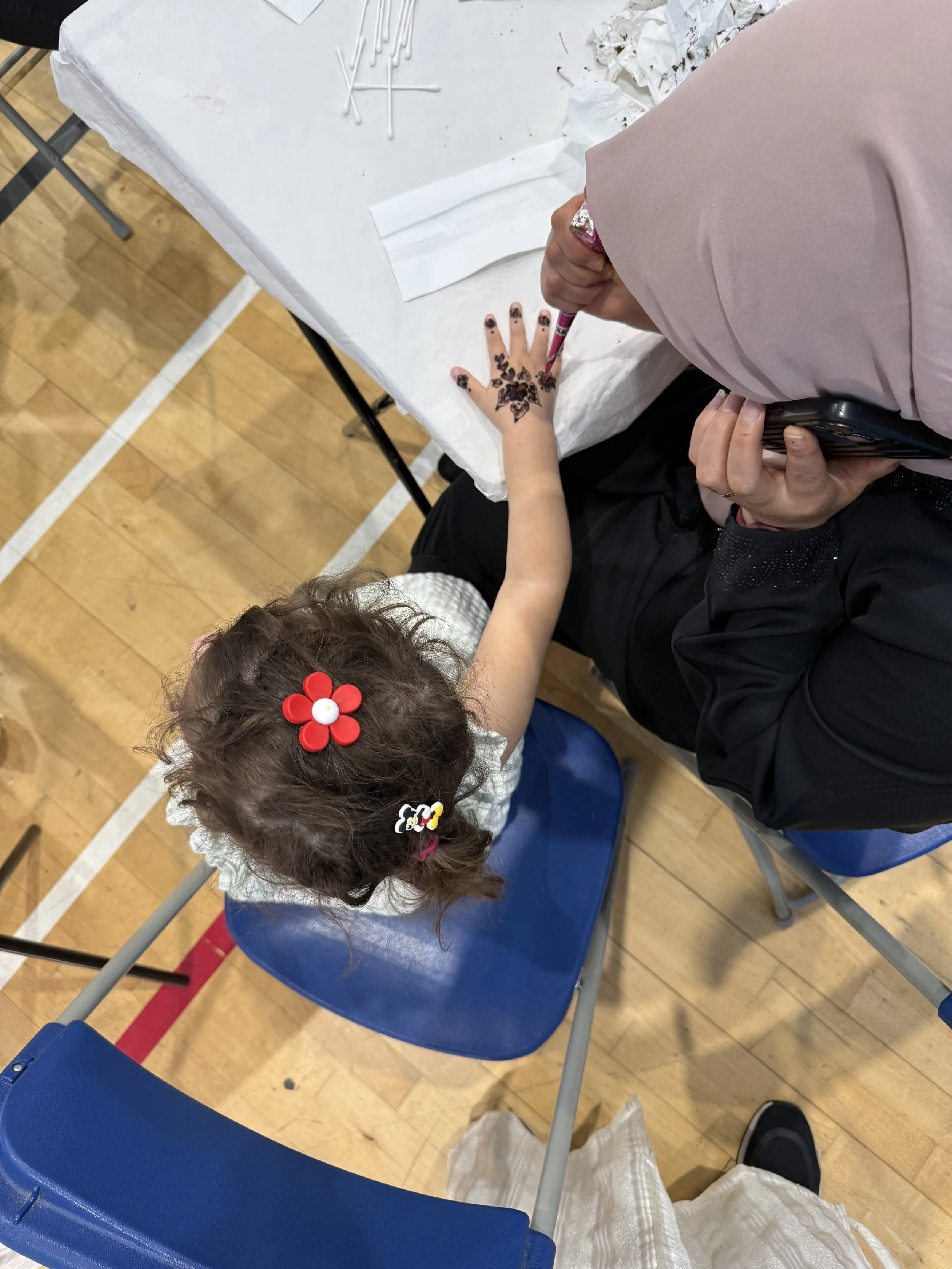 A person with henna tattoos on their hand is sitting at a table, having their hand decorated by another person, possibly a henna artist. The table has cotton swabs and tissues on it. The scene takes place in a gymnasium or similar indoor space with w