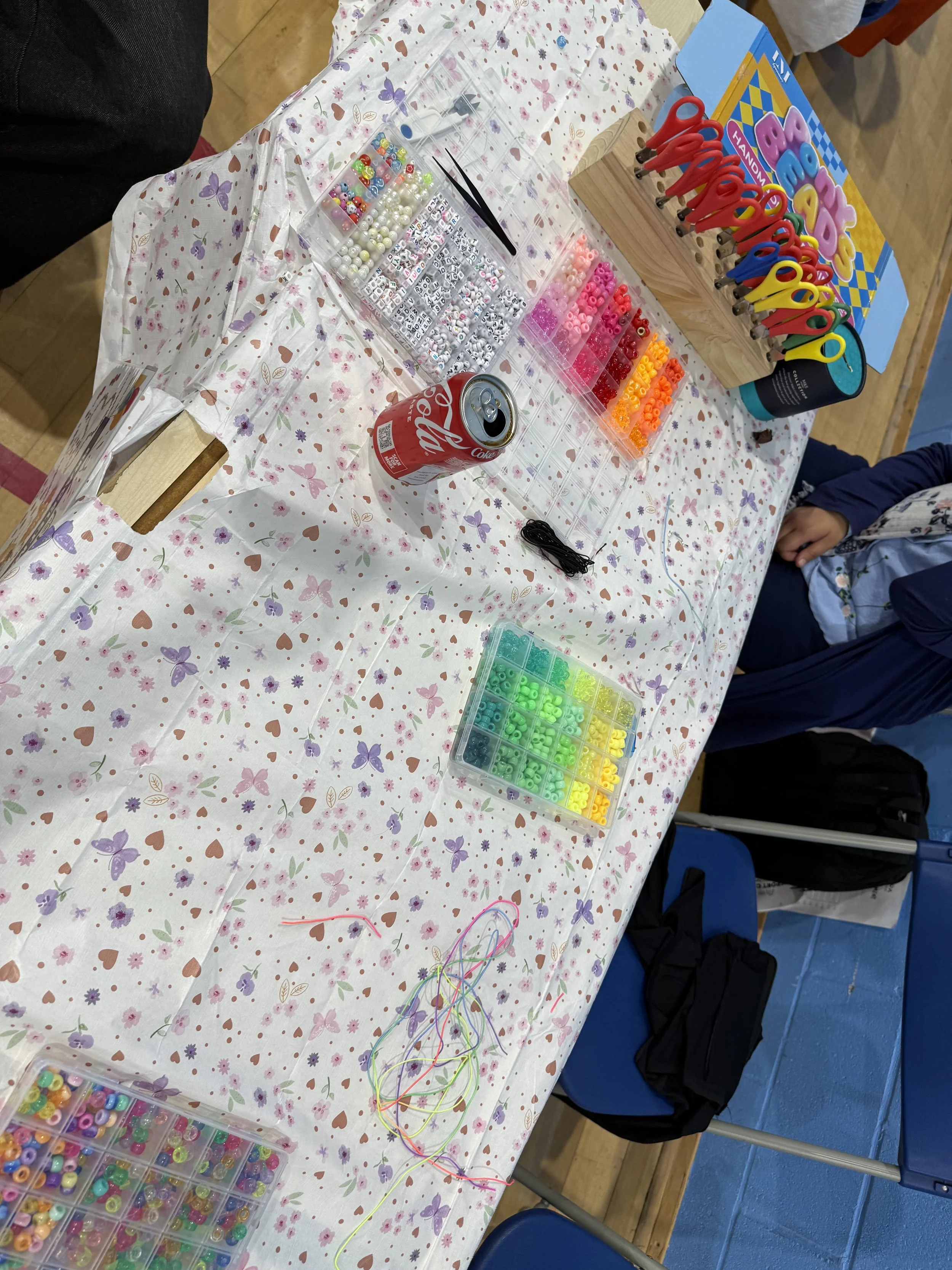 A craft table covered with a floral tablecloth displays various arts and crafts supplies, including colorful beads, scissors, thread, and a jewelry making kit. There are containers with small beads and a box of orange, pink, red, yellow, and green be