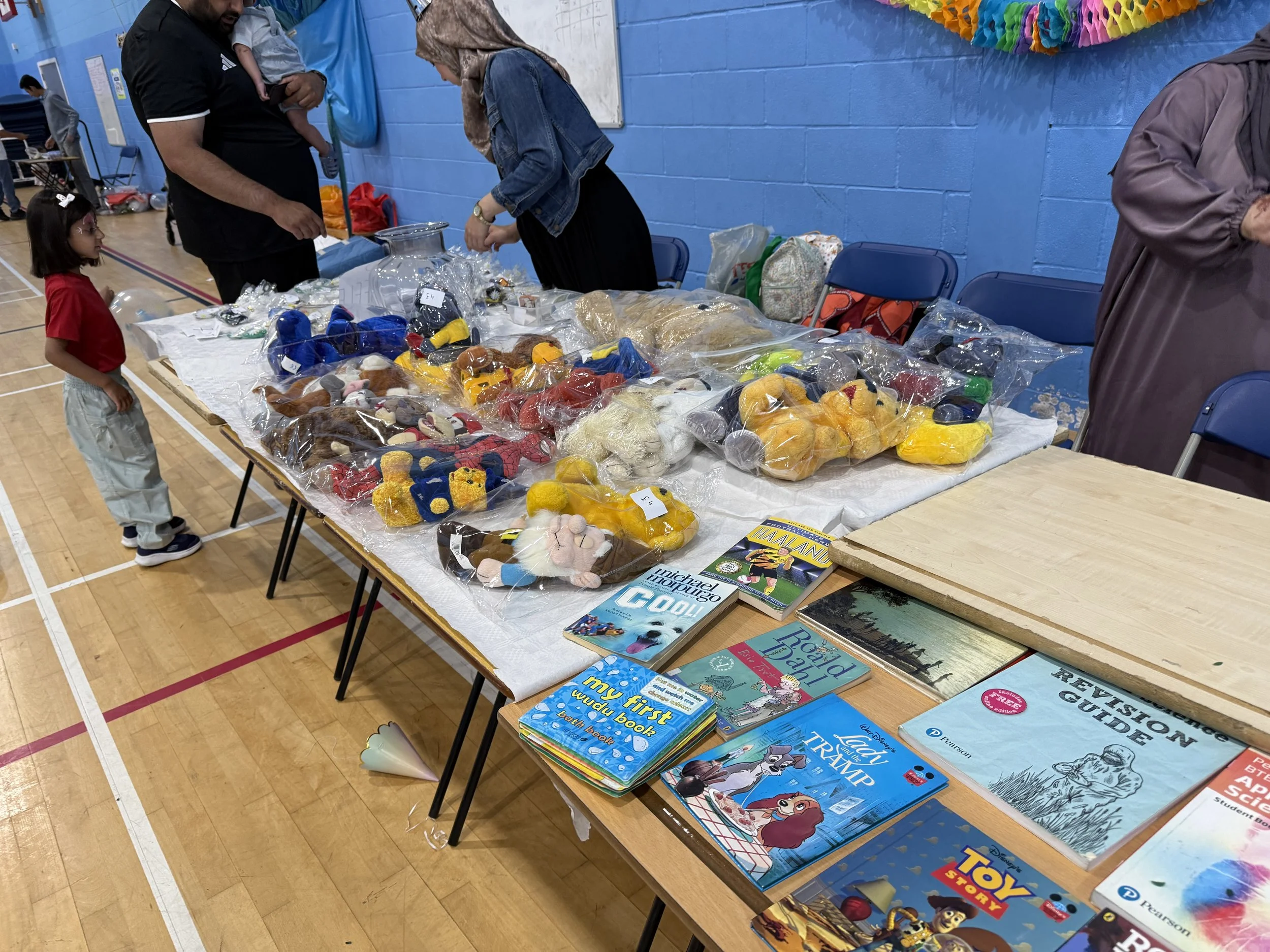 A table covered with toys and books, including stuffed animals, Barbie dolls, and children's books, in a gymnasium setting with blue walls and chairs in the background.