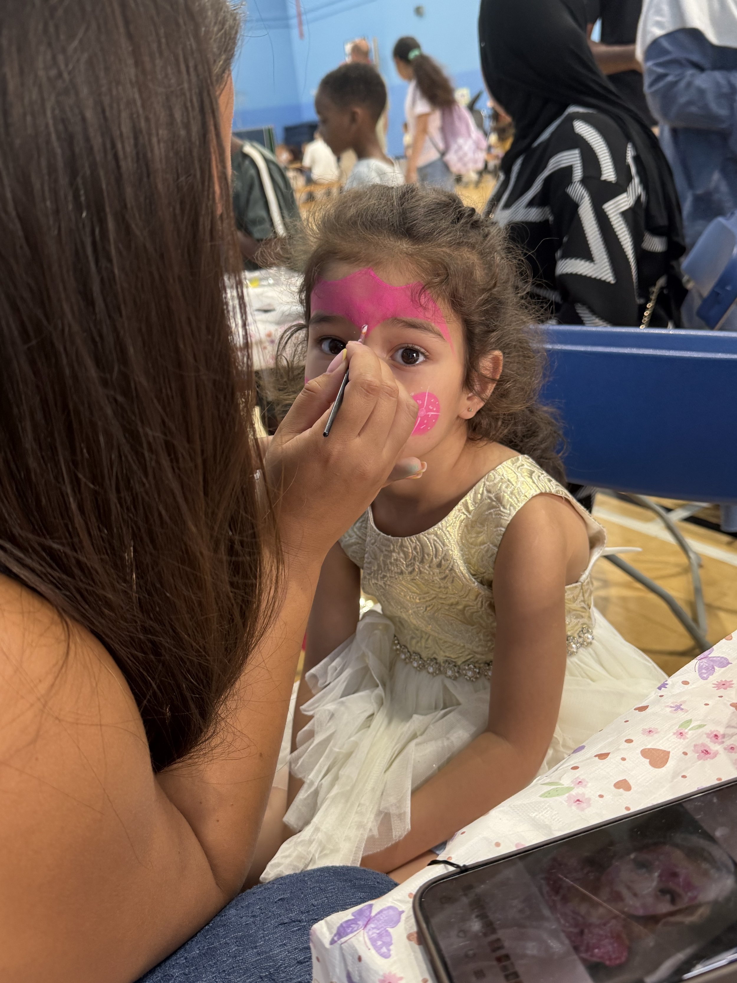 A young girl with dark curly hair and wearing a cream-colored dress is getting face paint applied by an adult woman with long brown hair. The girl has a pink butterfly design painted on her forehead and lips, and a pink drink is painted on her cheek.