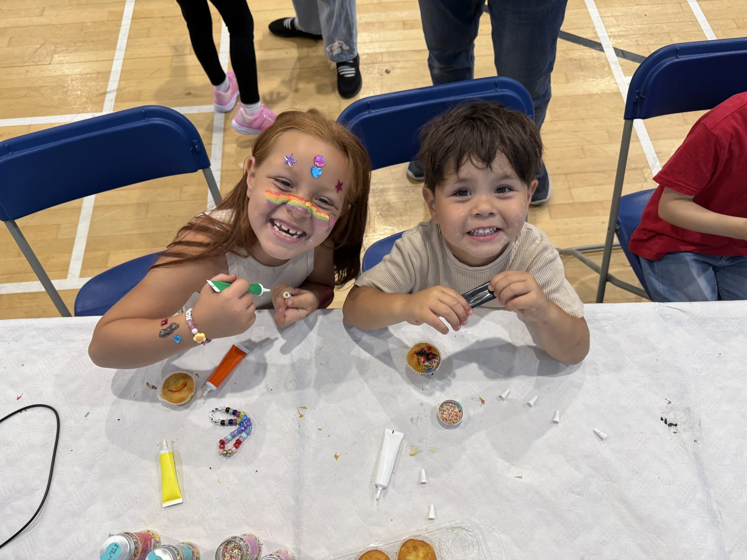 Two children sitting at a table decorated with colorful sprinkles, candies, and cupcake liners, smiling and enjoying a festive activity in an indoor gymnasium.