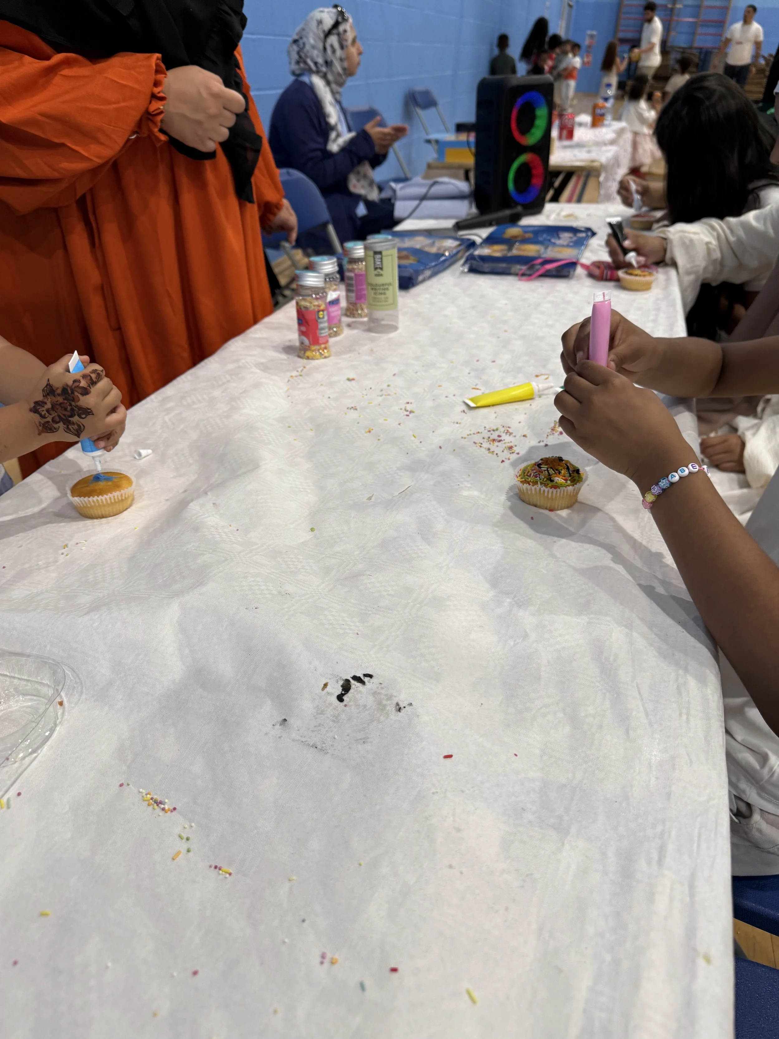 People gathered around a table decorating cupcakes with colorful sprinkles at an indoor event.