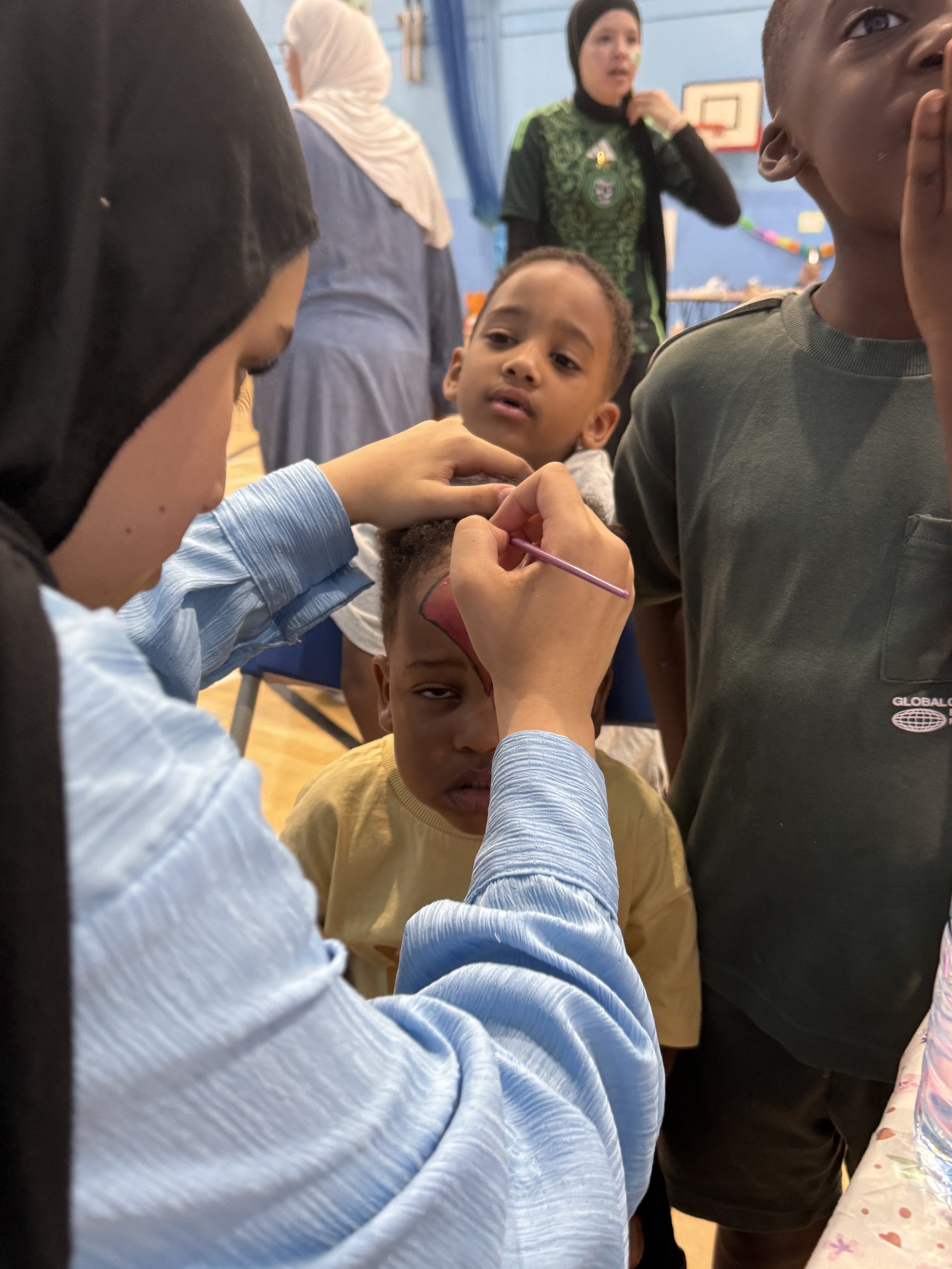 A young person with a painted face is being face painted by a woman wearing a blue shirt. Several children are gathered around watching, with a blue wall and a basketball hoop visible in the background.