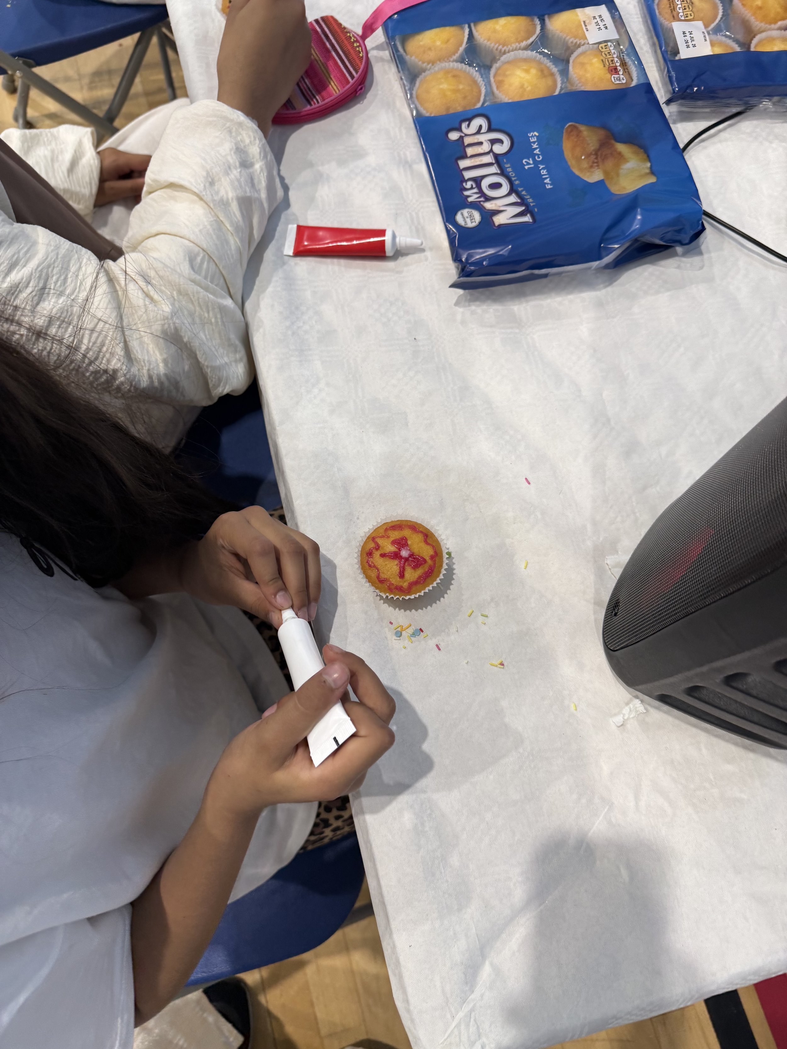 Person decorating a cupcake with pink icing from a tube at a table with a bag of muffins, a speaker, and a pink lunchbox.