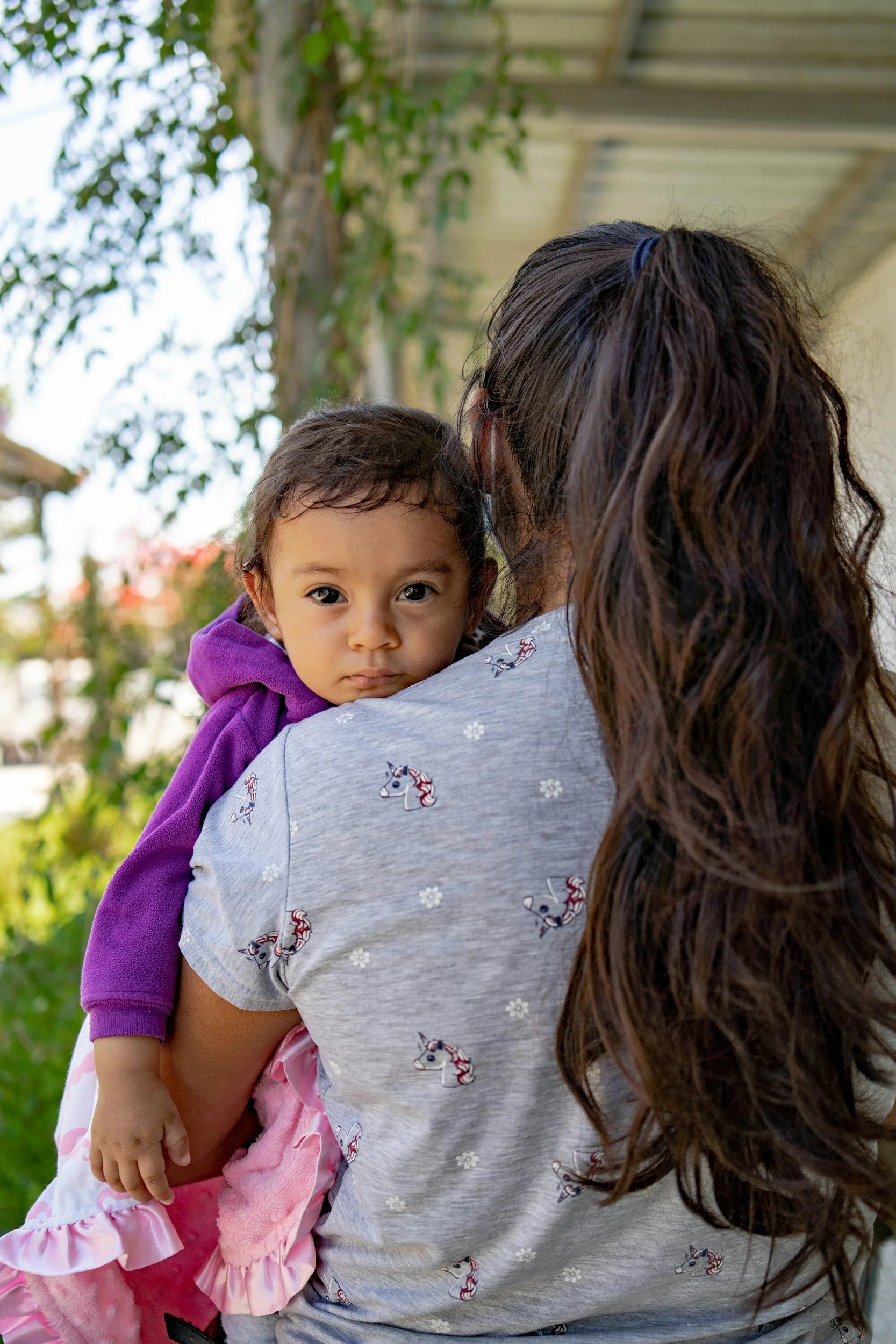 A woman holding a young girl outdoors, with trees and a building in the background. The girl is wearing a purple hoodie and looking at the camera.