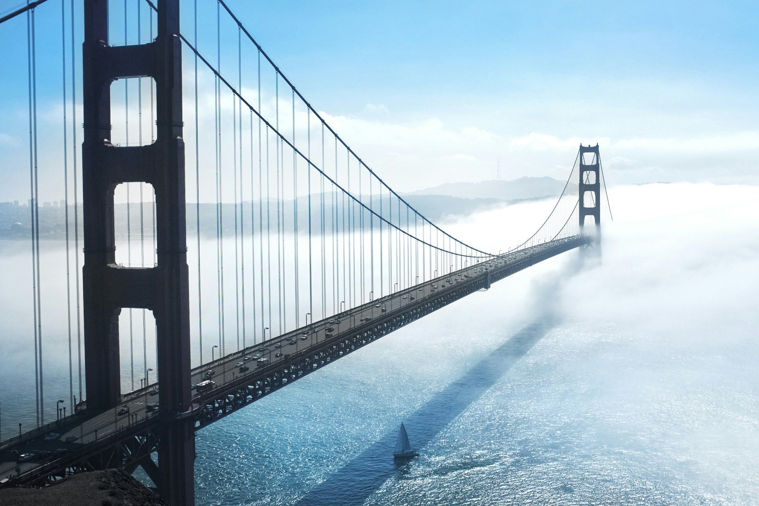 A view of the Golden Gate Bridge in San Francisco, California, partially covered by fog, with a sailboat in the water below and mountains in the background.