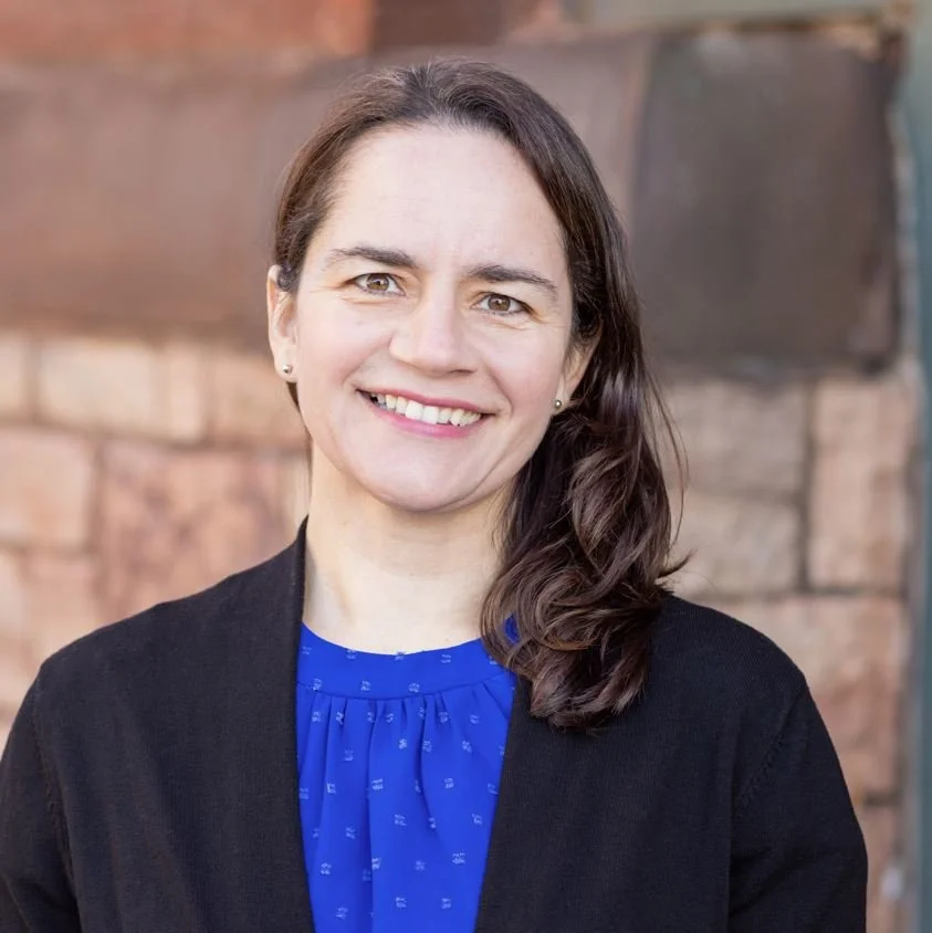 A smiling woman with long brown hair, wearing a black blazer over a blue blouse, stands outdoors in front of a brick wall.