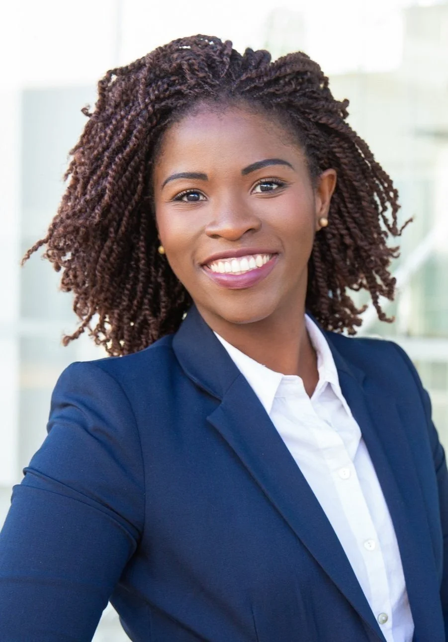 Portrait of a smiling woman wearing a blazer and white shirt, standing outdoors with a modern building in the background.