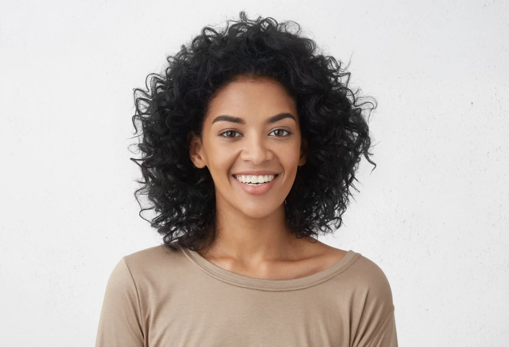 A smiling woman with curly black hair, wearing a beige top, standing against a plain white background.