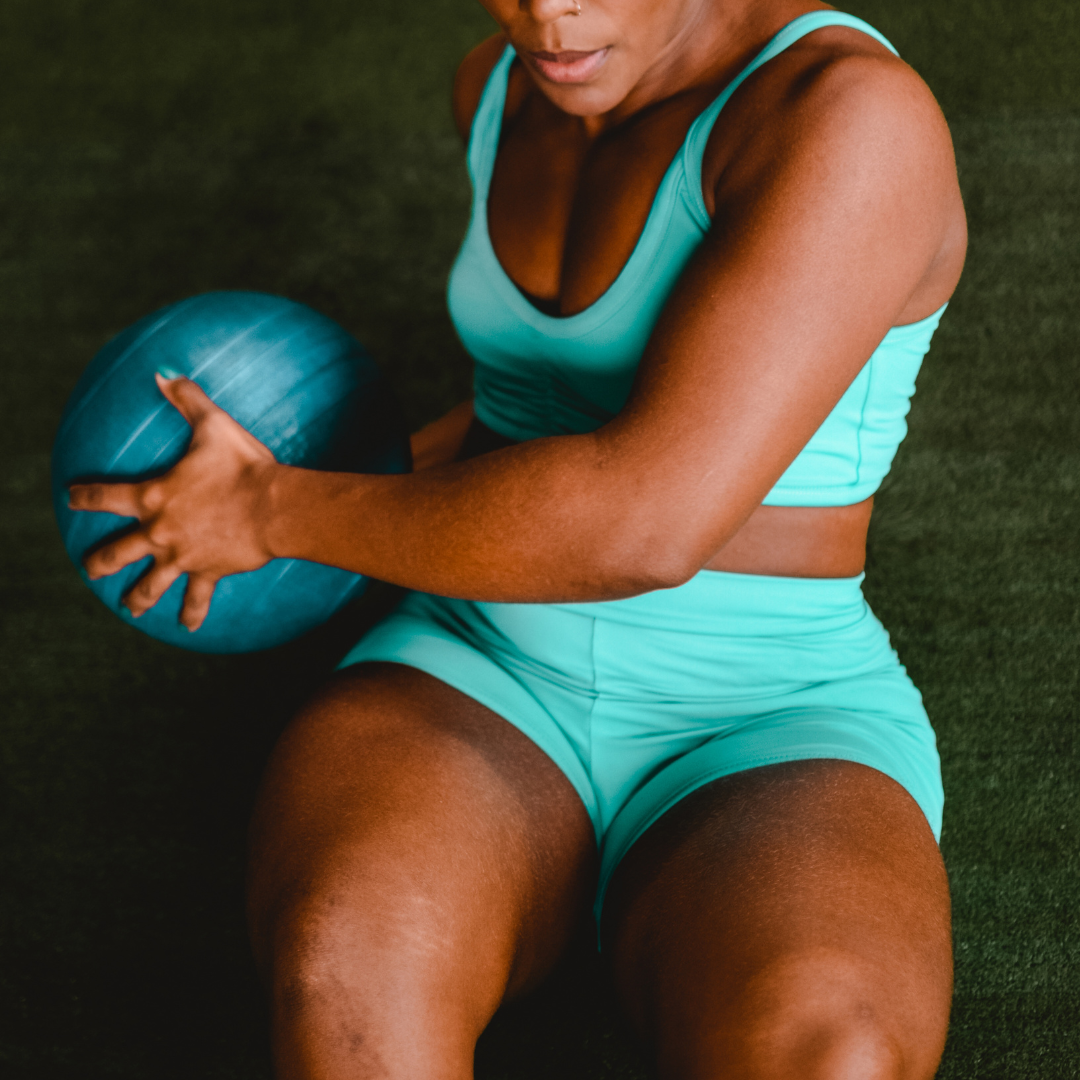 A woman in turquoise workout clothes sitting on a gym mat, holding a blue exercise ball.
