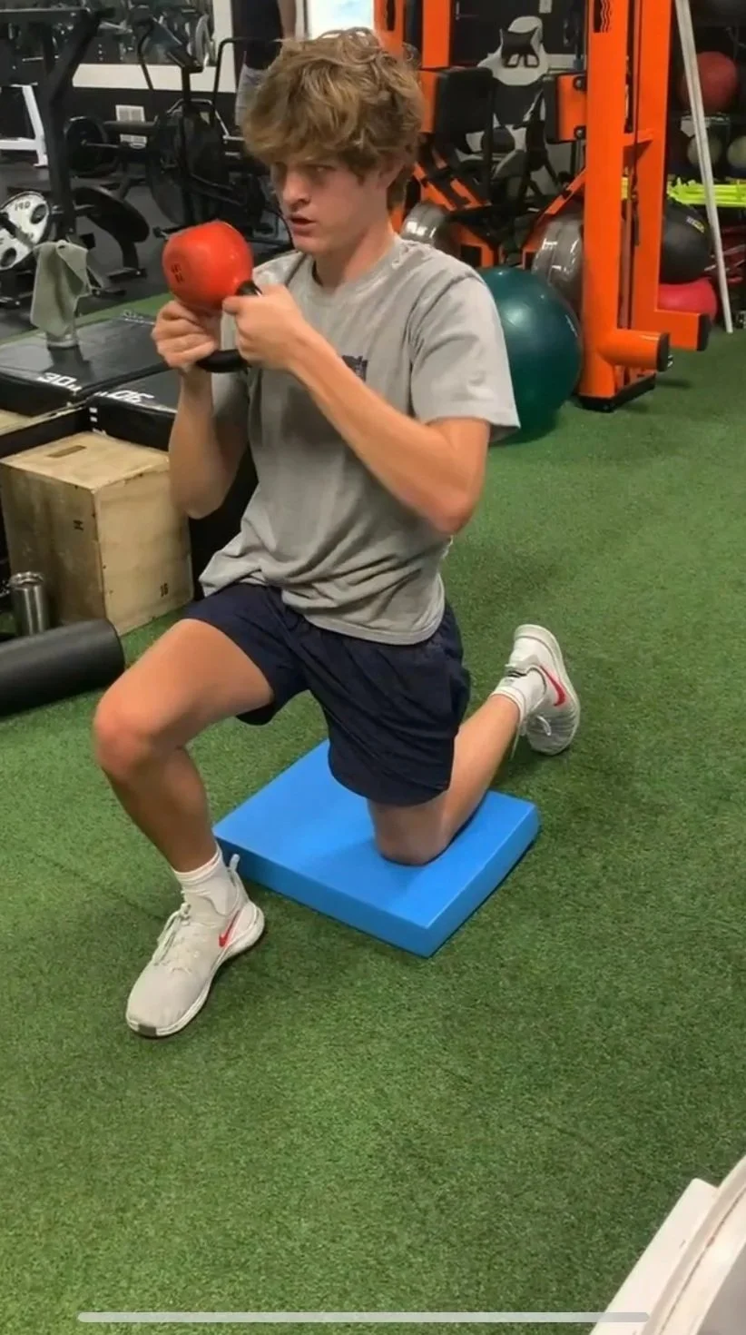 A young man kneeling on one knee on an exercise mat in a gym, holding a red kettlebell close to his chest, surrounded by exercise equipment and balls.
