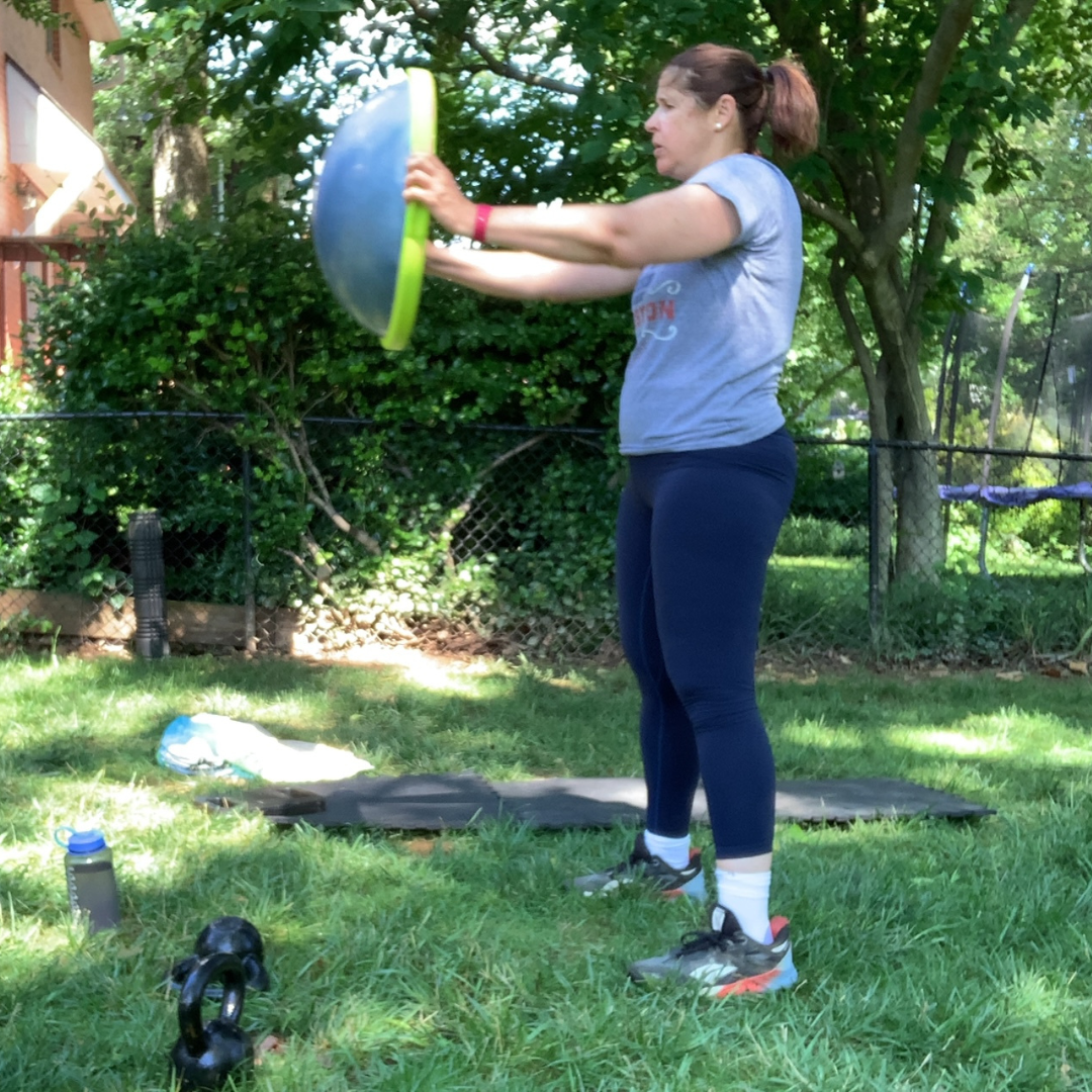 Woman exercising outdoors holding a weight plate in front of her with greenery and a fence in the background.