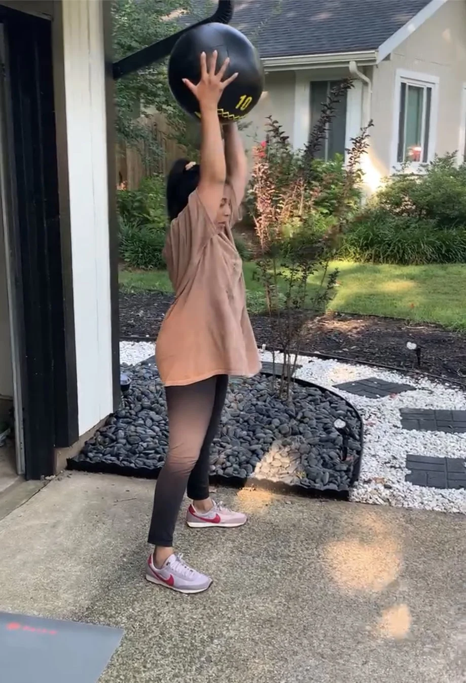 A girl in a peach-colored shirt and black leggings is lifting a black medicine ball with yellow markings above her head outdoors on a concrete patio, with a garden and house in the background.