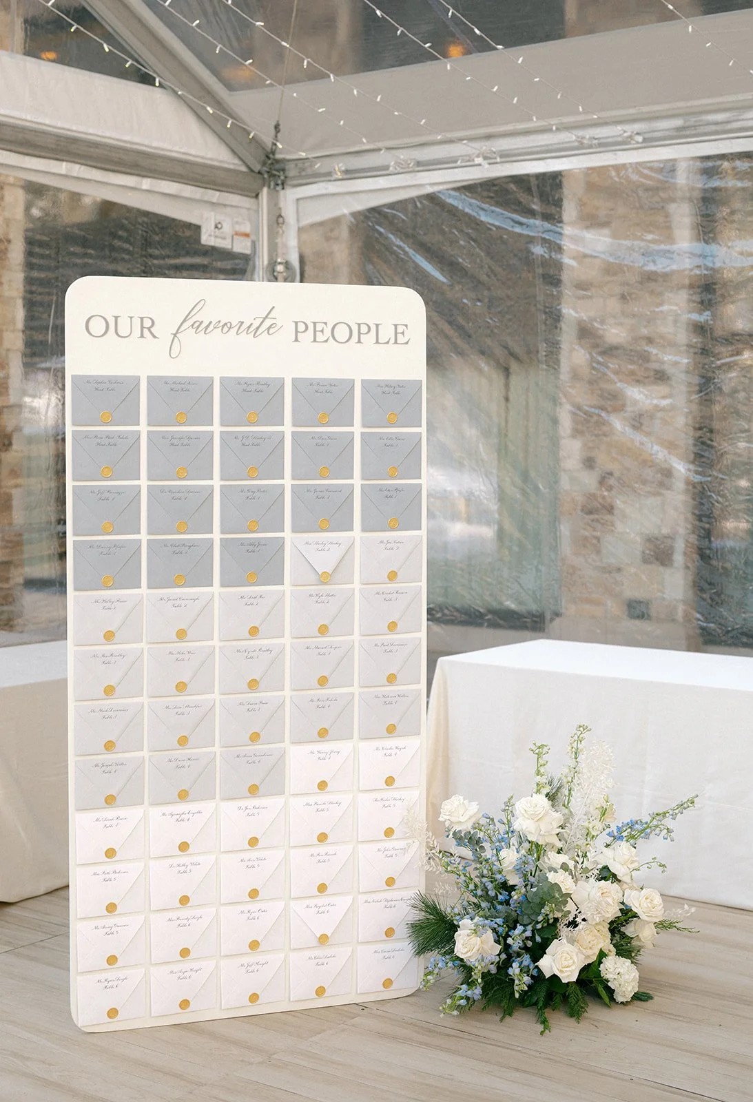  Winter wedding escort card display with gray and white envelopes sealed in gold wax, styled with white and blue floral arrangement. 