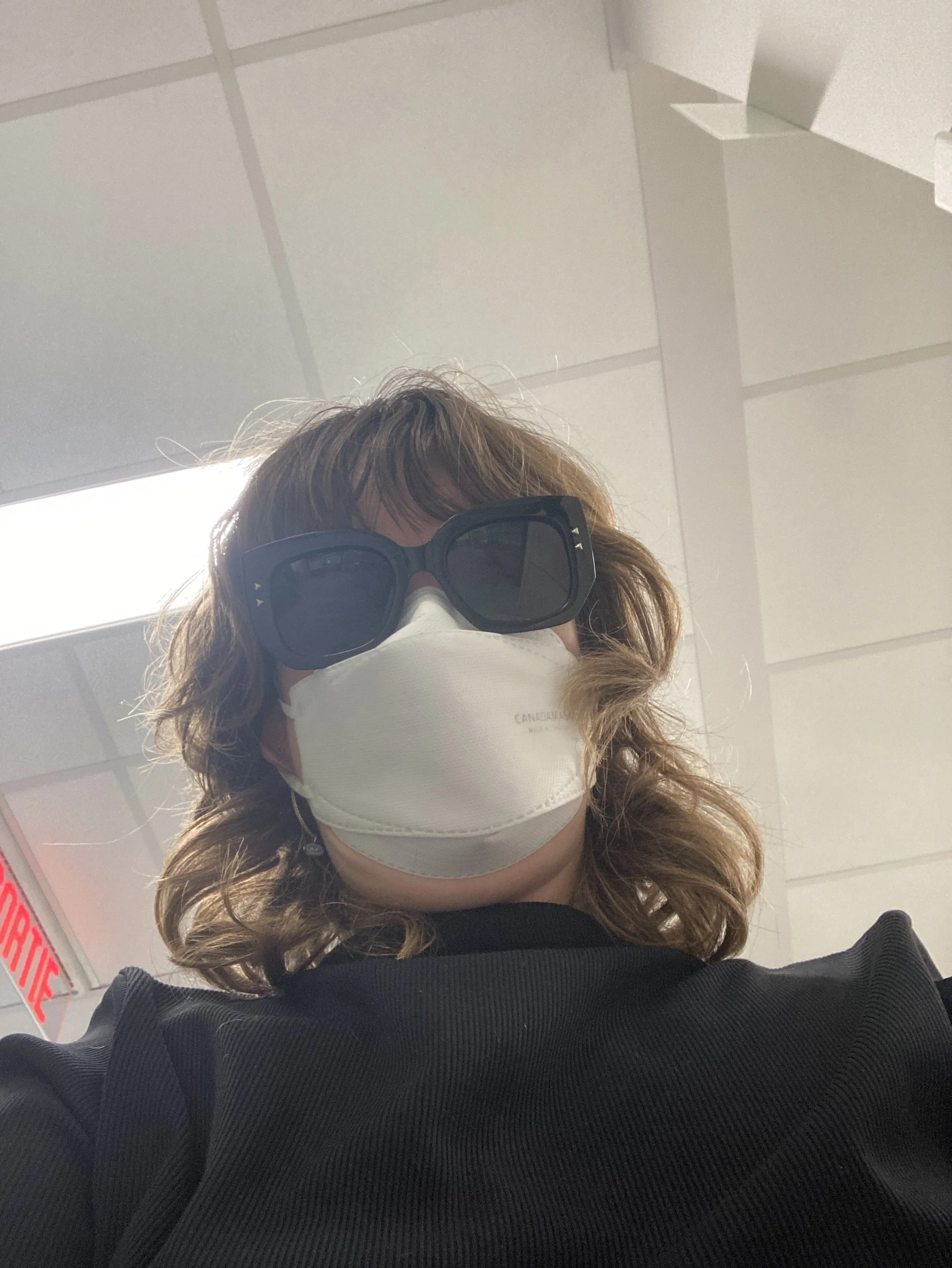 A selfie of Aimee Louw, a white woman with light brown hair, wearing big black sunglasses and a white KN95 mask. Above her are the white ceiling tiles of the hospital and an exit sign.