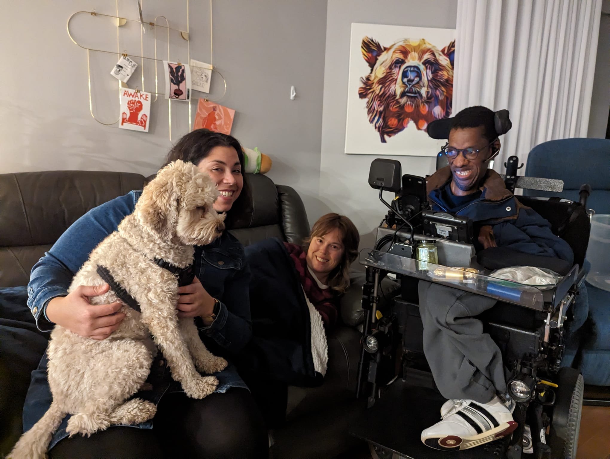 A group photo of three friends and one dog in a living room. The dog is seated on a woman’s lap and next to her on a gray couch is Aimee Louw lying down on the couch under a blanket smiling.  Beside them is their friend in his motorized wheelchair.