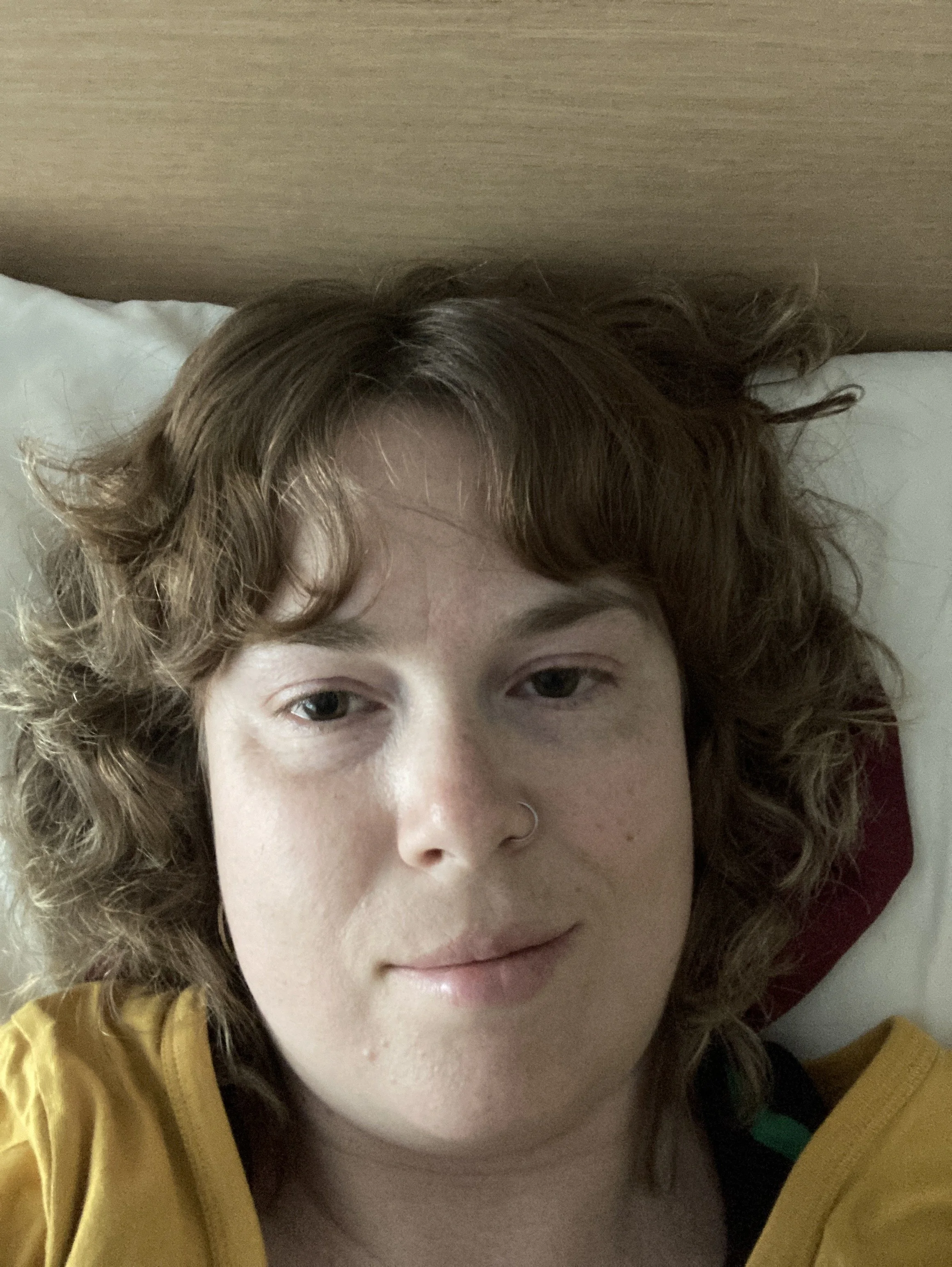 A selfie of Aimee Louw, lying down in bed, wearing a yellow shirt and smiling loosely at the camera with slightly squinted eyes and an exhausted pale face.